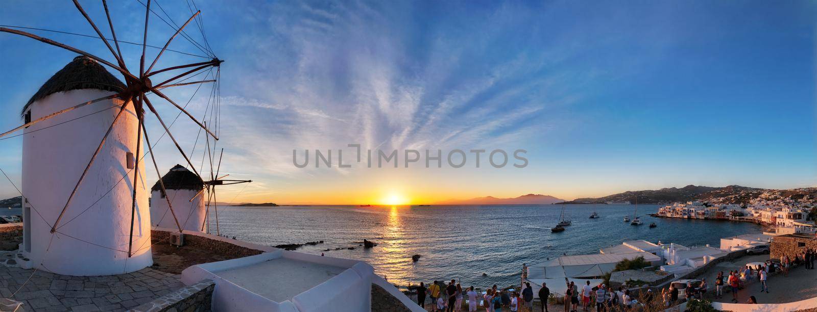 MYKONOS, GREECE - MAY 29, 2019: Panorama of traditional greek windmills on Mykonos island on sunset with dramatic sky and Little Venice quarter with tourist crowd