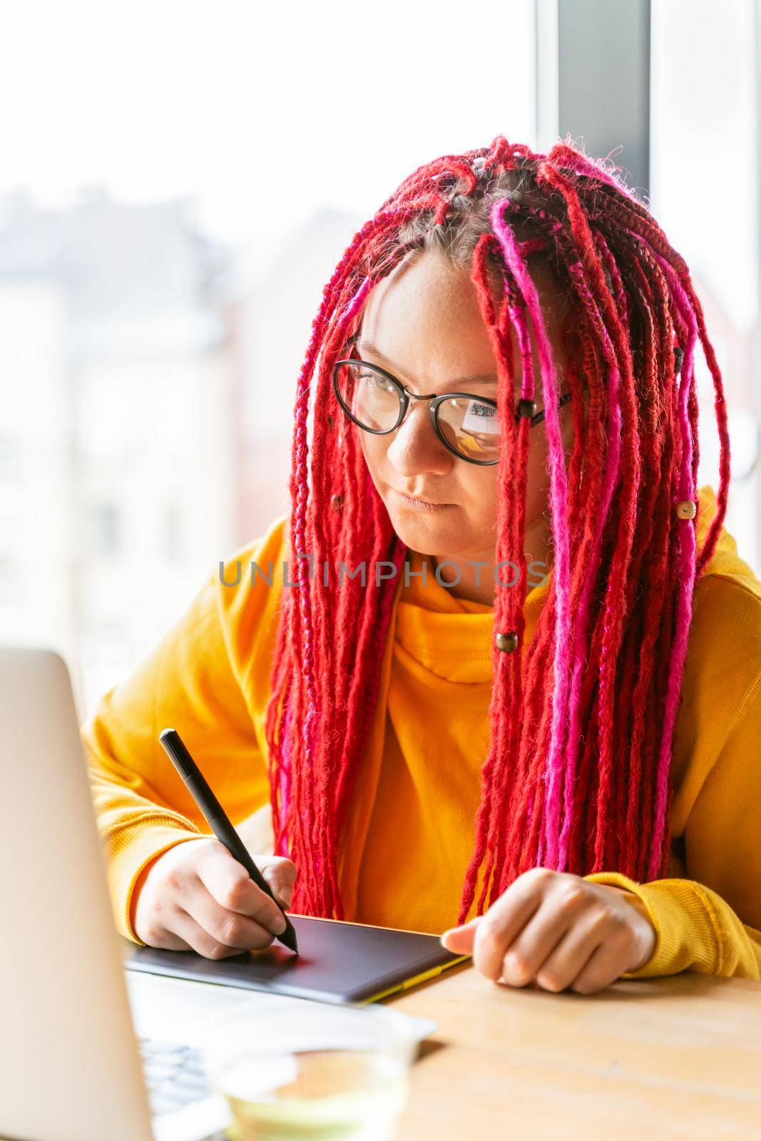 Girl designer, illustrator draws on a tablet, looks into a laptop. The concept of the remote work of a creative hobby, freelance. Woman with long pink hair, bright appearance, vertical