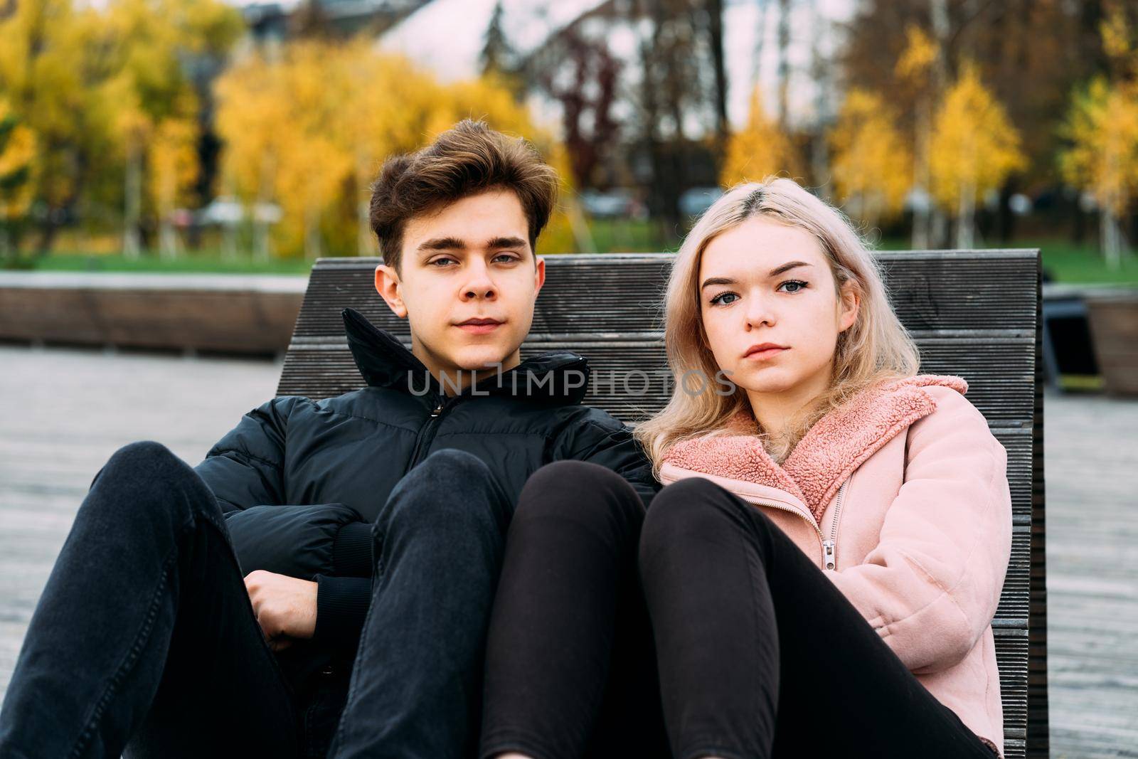 Loving teenagers are sitting on park bench in autumn, looking straight ahead. Teenage love concept. Outdoor portrait of cute brunette guy in dark jacket, beautiful young blonde girl in light pink jacket.