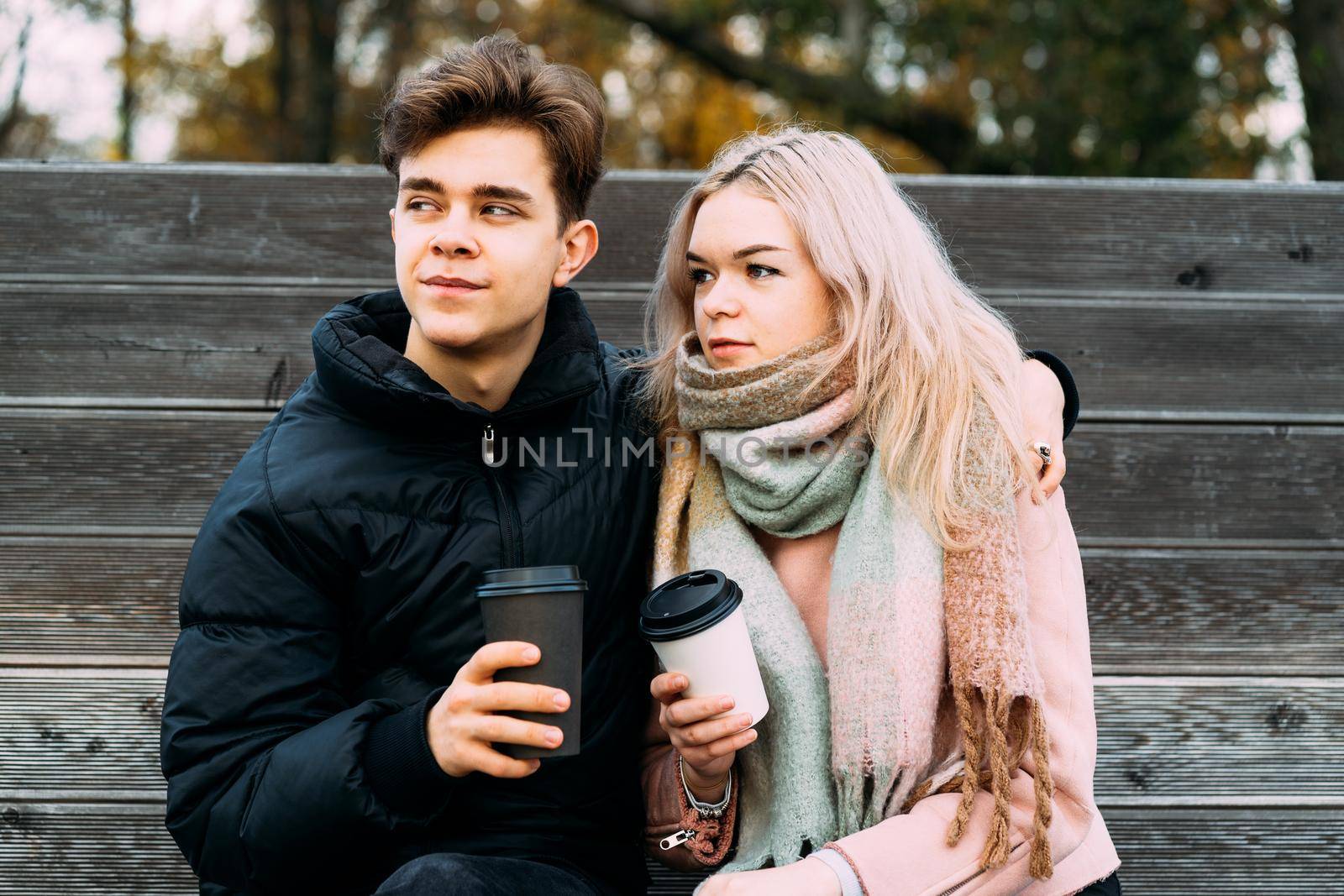 Lovely brunette guy and pretty girl blonde drink coffee and chat on a date, dreaming. Loving teenagers are sitting on park bench. Teen Love Concept. They are happy, smiling, laughing