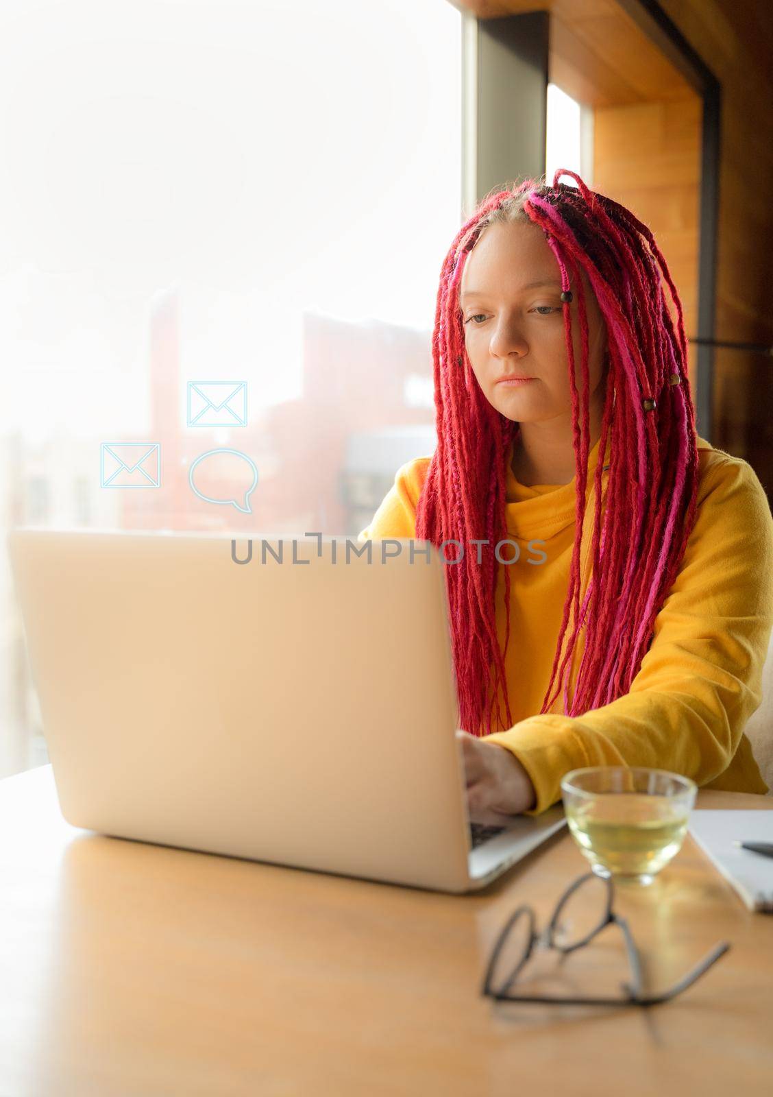 Digital nomad concept. Girl freelancer remotely working on a laptop in a cafe, coworking. Woman with long pink dreadlocks in informal setting, in casual comfortable clothes sitting at table, vertical