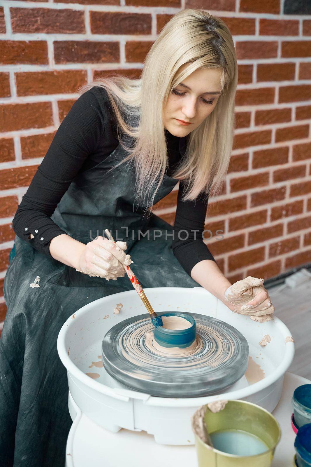 Happy woman making ceramic pottery on wheel, paints blue. Concept for woman in freelance, business. Handcraft product. Earn extra money, side hustle, turning hobbies into cash and passion into a job
