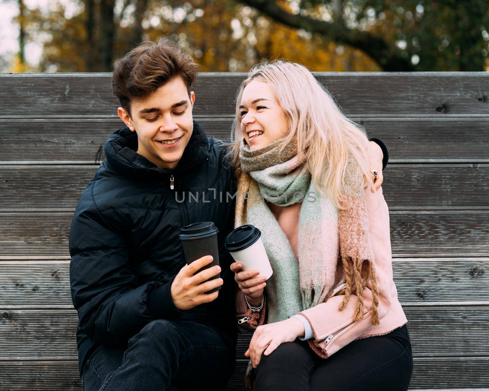 Lovely brunette guy and pretty girl blonde drink coffee and chat on date, laugh. Loving teenagers are sitting on park bench. Teen Love Concept. They are happy, smiling, laughing. Evening