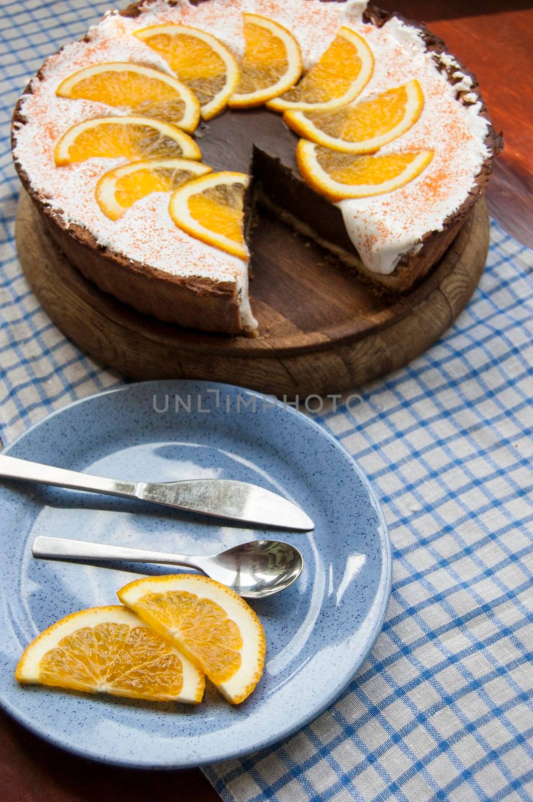 Chocolate cheesecake on a wooden board. Near blue plate, knife and teaspoon adn two slice of orandge