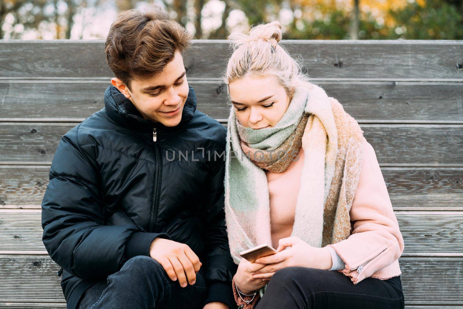 Loving teenagers on date look at mobile phones, girl shows guy something interesting on a mobile phone. Immersion in virtual world, social networks. Concept of teen love, Smombie dating