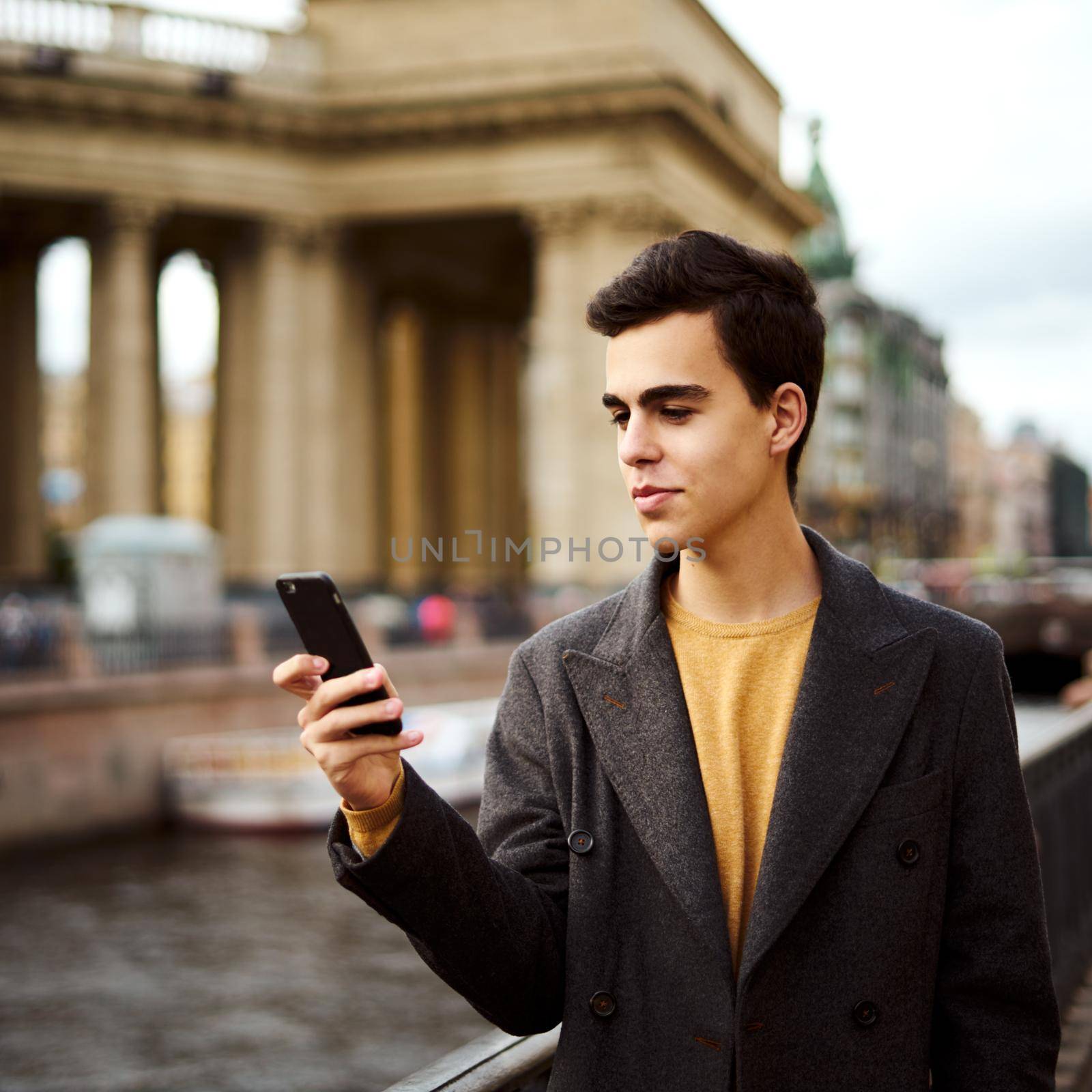Handsome stylish fashionable man talking on phone, dialing chat message, brunette in elegant gray coat is standing on street in a historical center. Young man with dark hair, thick eyebrows.