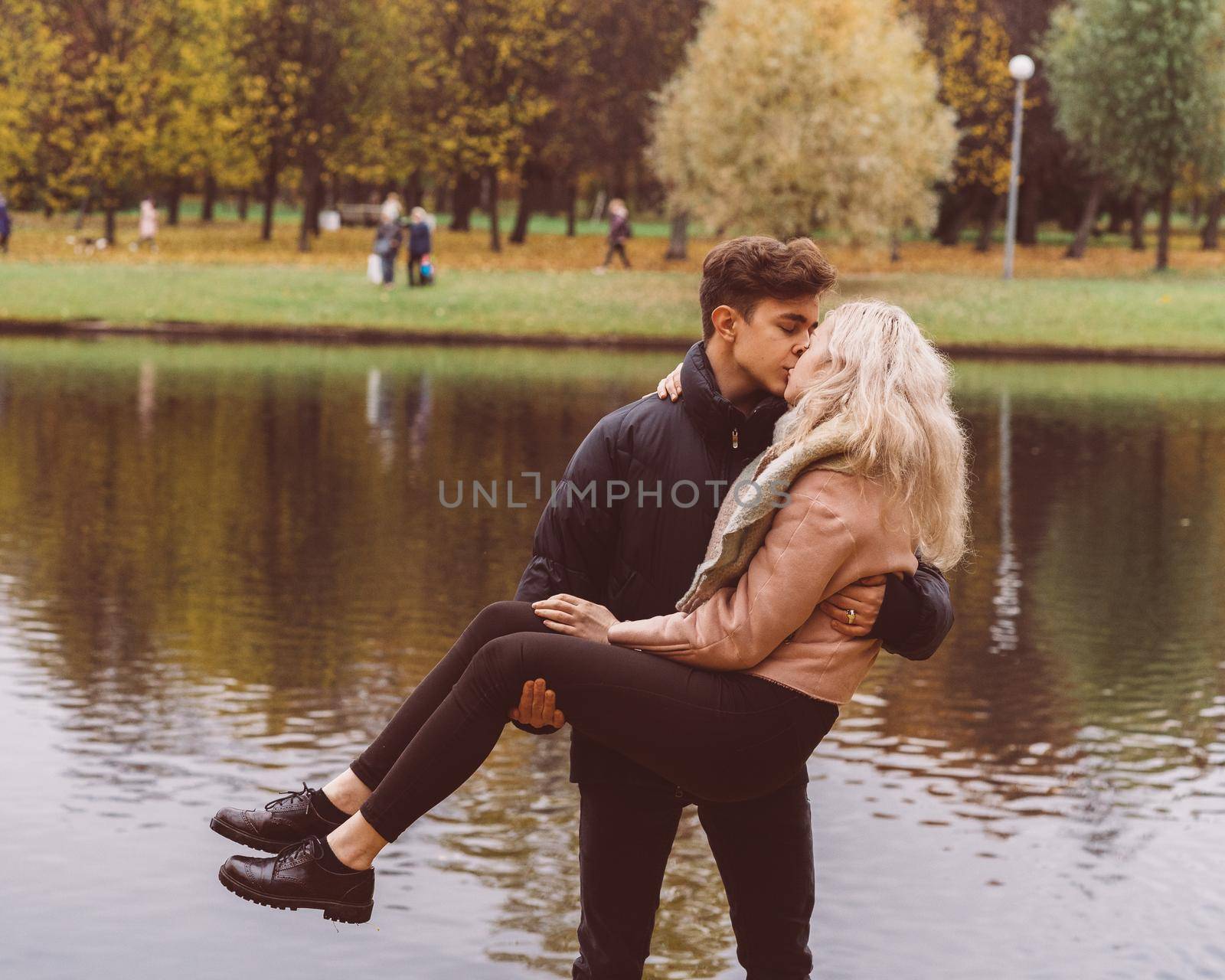 Cute brunette guy holds in his arms and kisses a beautiful blonde girl. Loving teens are happy, smiling, kissing. The concept of teenage love walking outdoors