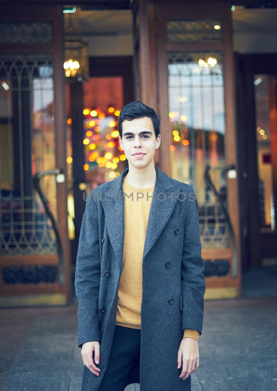 Handsome stylish fashionable man, brunette in elegant gray coat, stands on the street in historical center of St. Petersburg. Young man with dark hair, thick eyebrows, vertical