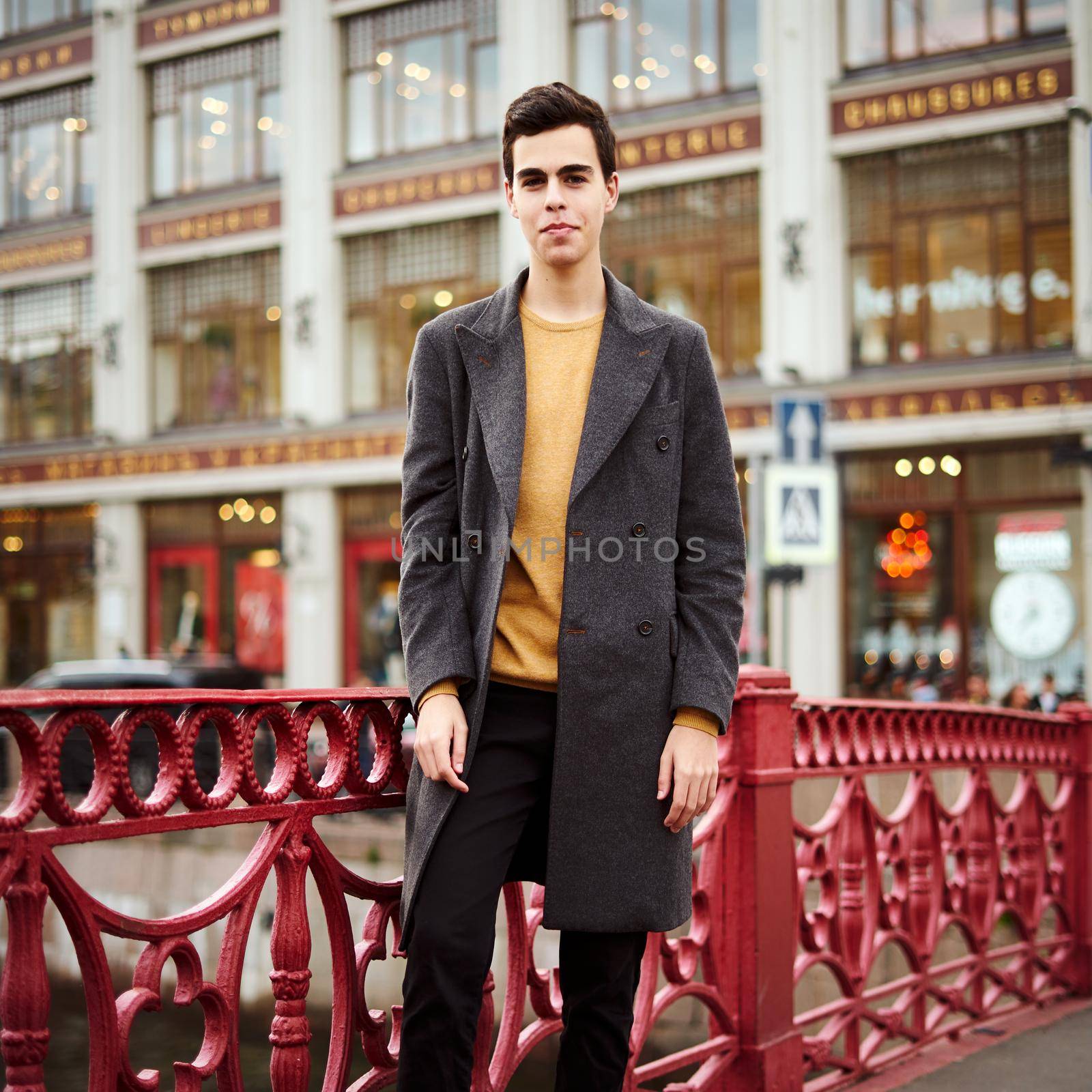 Handsome stylish fashionable man, brunette in elegant gray coat, stands on the street in historical center of St. Petersburg. Young man with dark hair, thick eyebrows.