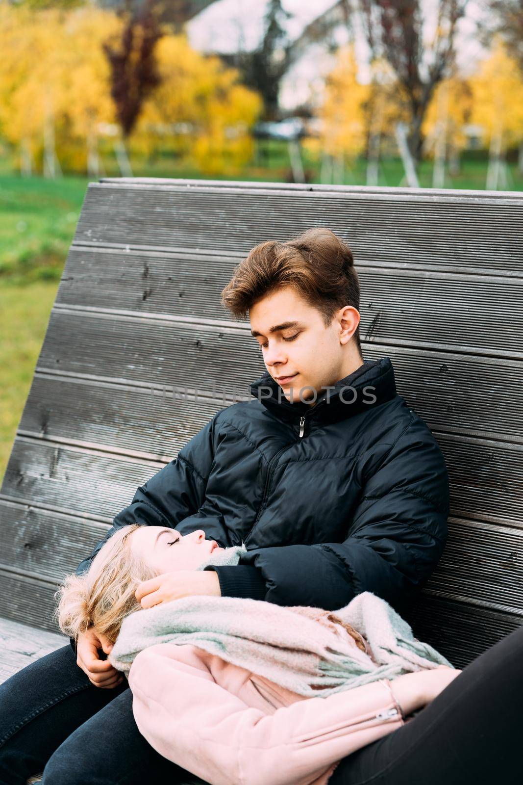 Teenagers in love are sitting on park bench in autumn, chatting cheerfully, talking. Cute blonde girl lies, her head on boy lap. Teenage love concept, vertical