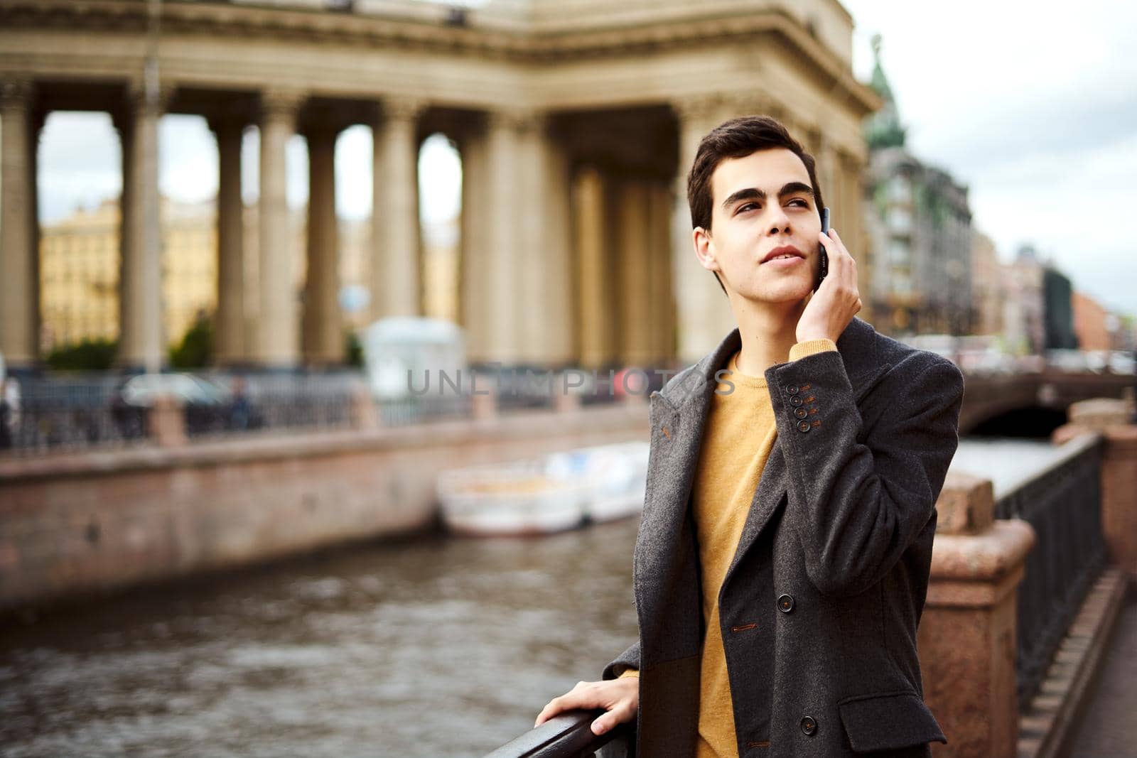 Handsome stylish fashionable man is talking on phone, brunette in en elegant gray coat is standing on street in historical center of St. Petersburg. Young man with dark hair, thick eyebrows.