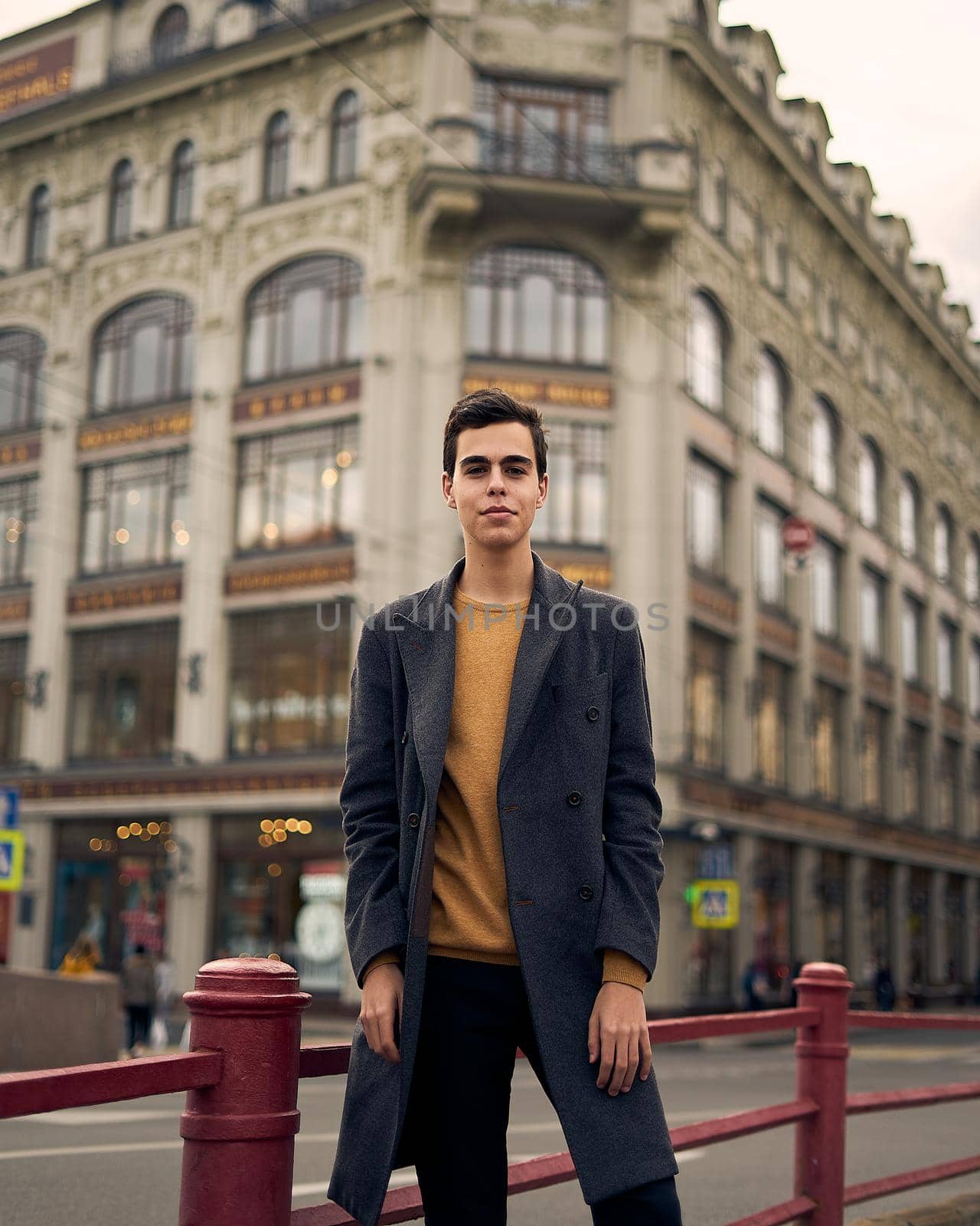 Handsome stylish fashionable man, brunette in elegant gray coat, stands on street in historical center of St. Petersburg. Young man with dark hair, thick eyebrows. by NataBene