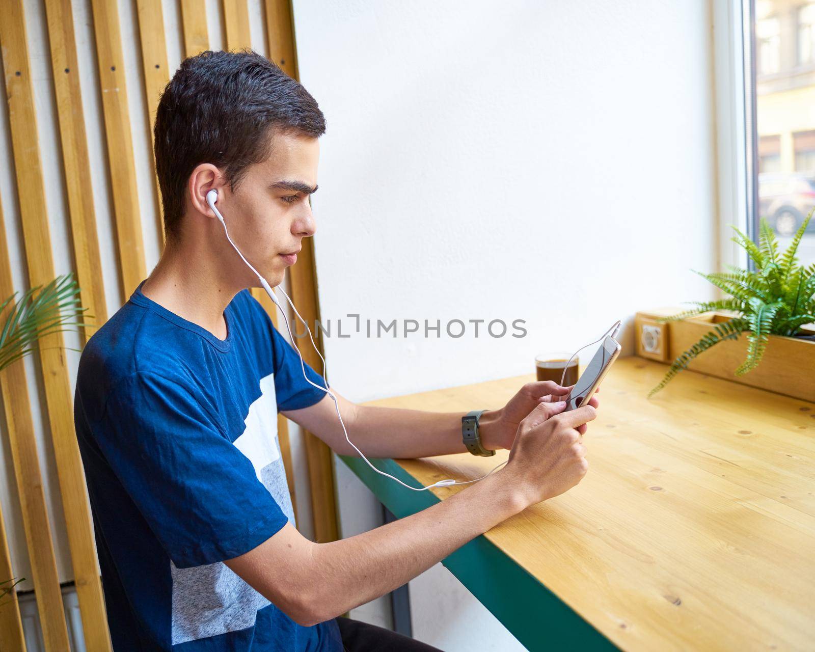 Young adult texting message on smart phone and listening to music on earphones in coffee shop, copy space