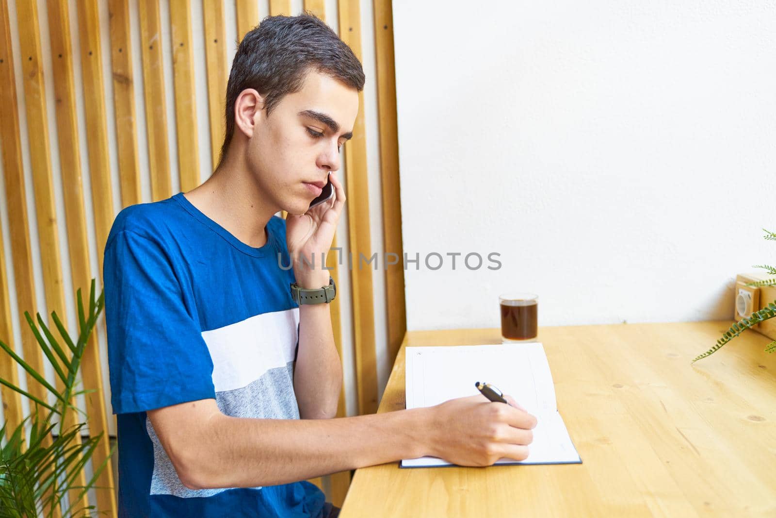 Young man writing information to notepad and speaking mobile phone, teenager in casual clothes plans a schedule, copy space