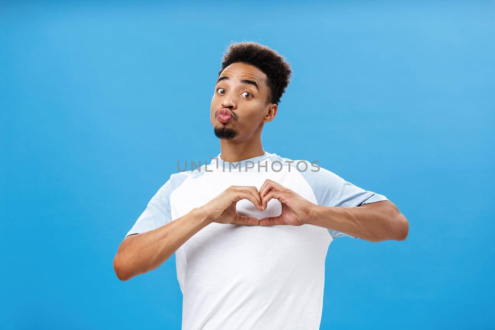 Funny and cute dark-skinned young male student in t-shirt folding lips in mwah or kiss showing heart gesture over breast giving love and affection signs to girl he likes over blue wall. Emotions concept