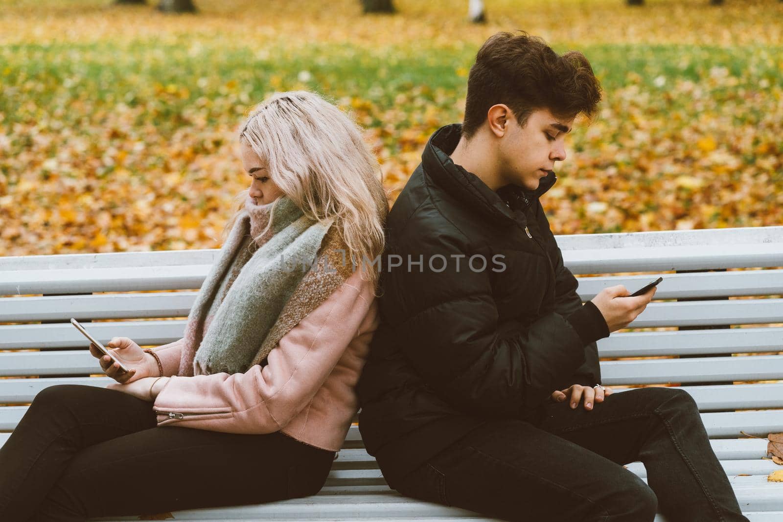 Loving teenagers on date look at mobile phones, sit on park bench in the fall. Immersion in virtual world, social networks. Concept of teenage love, Smombie. Teen dating, top view