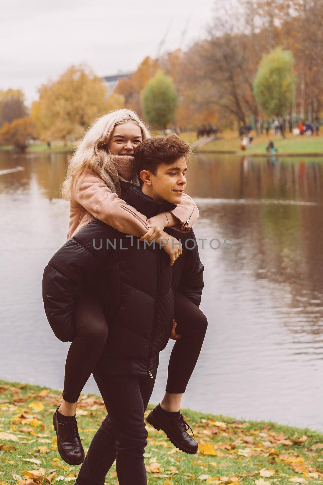 A pretty blonde girl is sitting on back of a charming brunette man. Loving teens are happy, smiling, looking away. Concept of teenage love walking outdoors