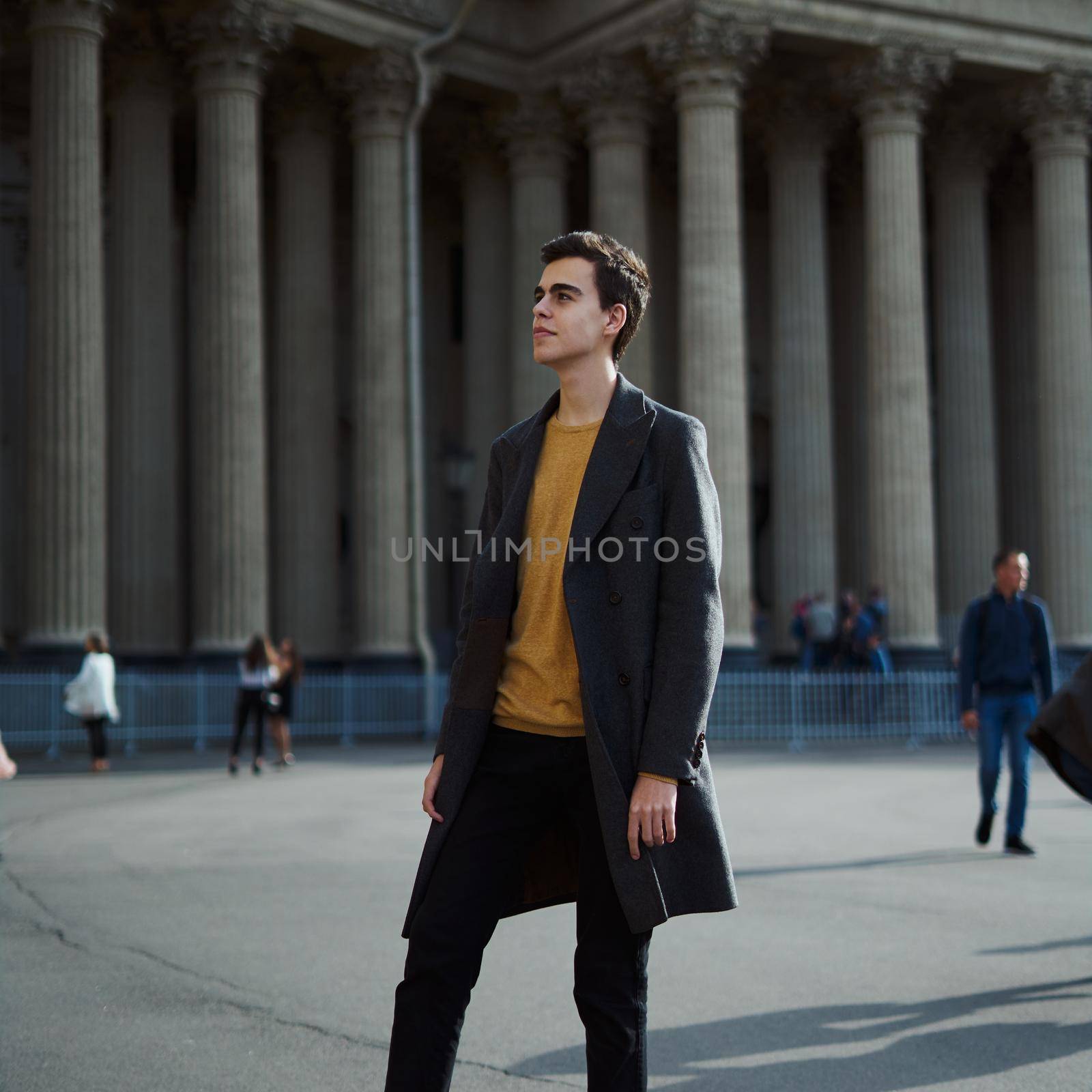 Handsome stylish fashionable man, brunette in an elegant gray coat, stands on street in historical center of St. Petersburg. Young man with dark hair, thick eyebrows.