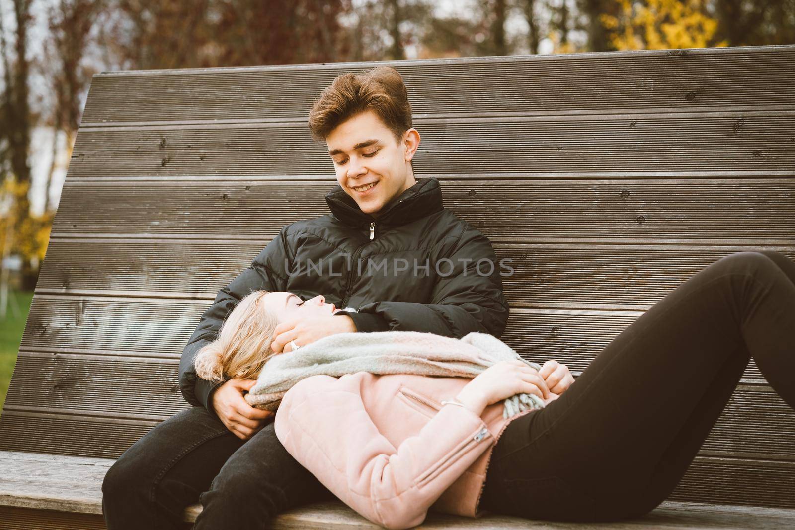 Teenagers in love are sitting on park bench in a autumn, chatting cheerfully, talking. Cute blonde girl lies, her head on boy lap, guy smiles. Teenage love concept.