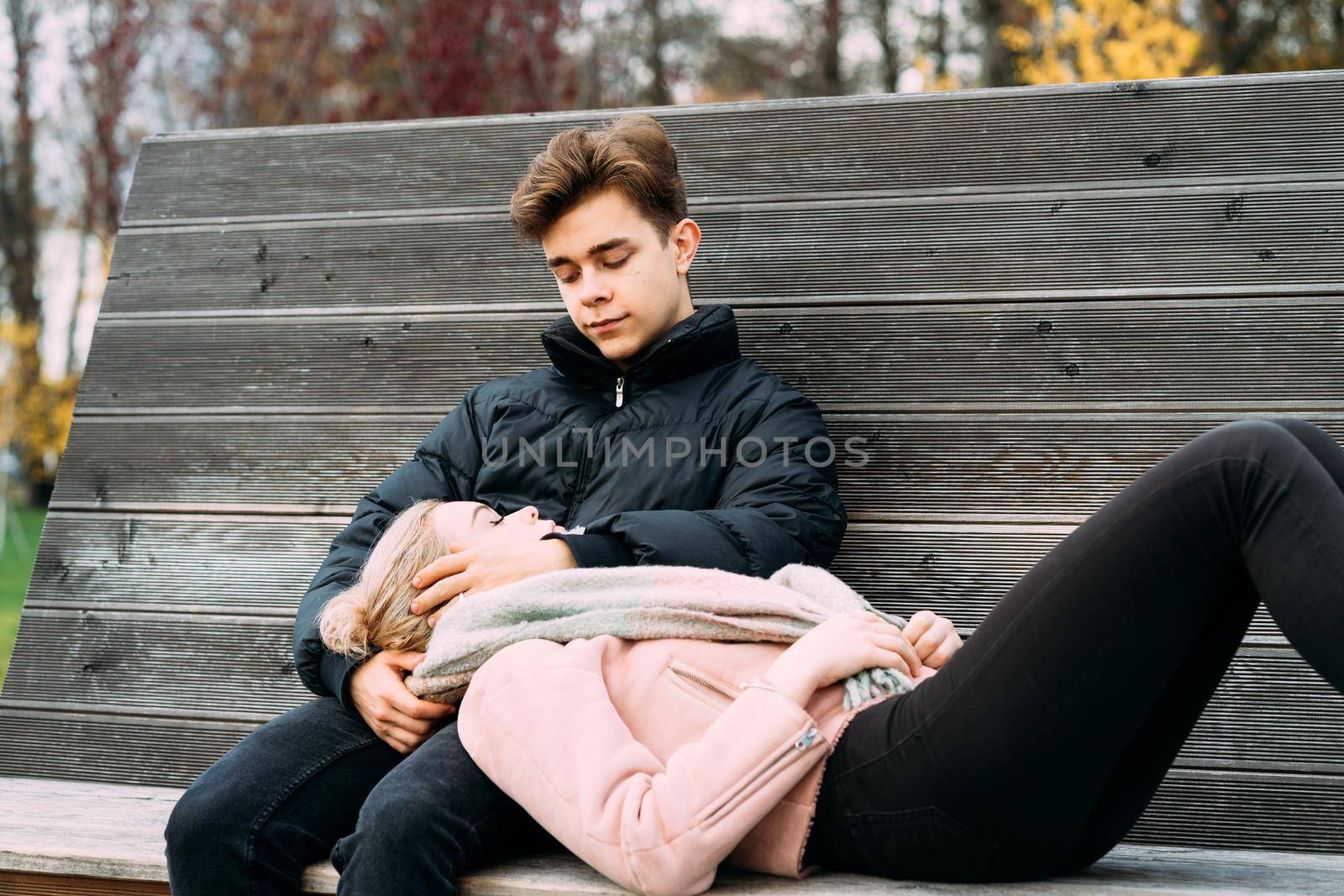 Teenagers in love are sitting on park bench in a autumn, chatting cheerfully, talking. Cute blonde girl lies, her head on boy lap. Teenage love concept.