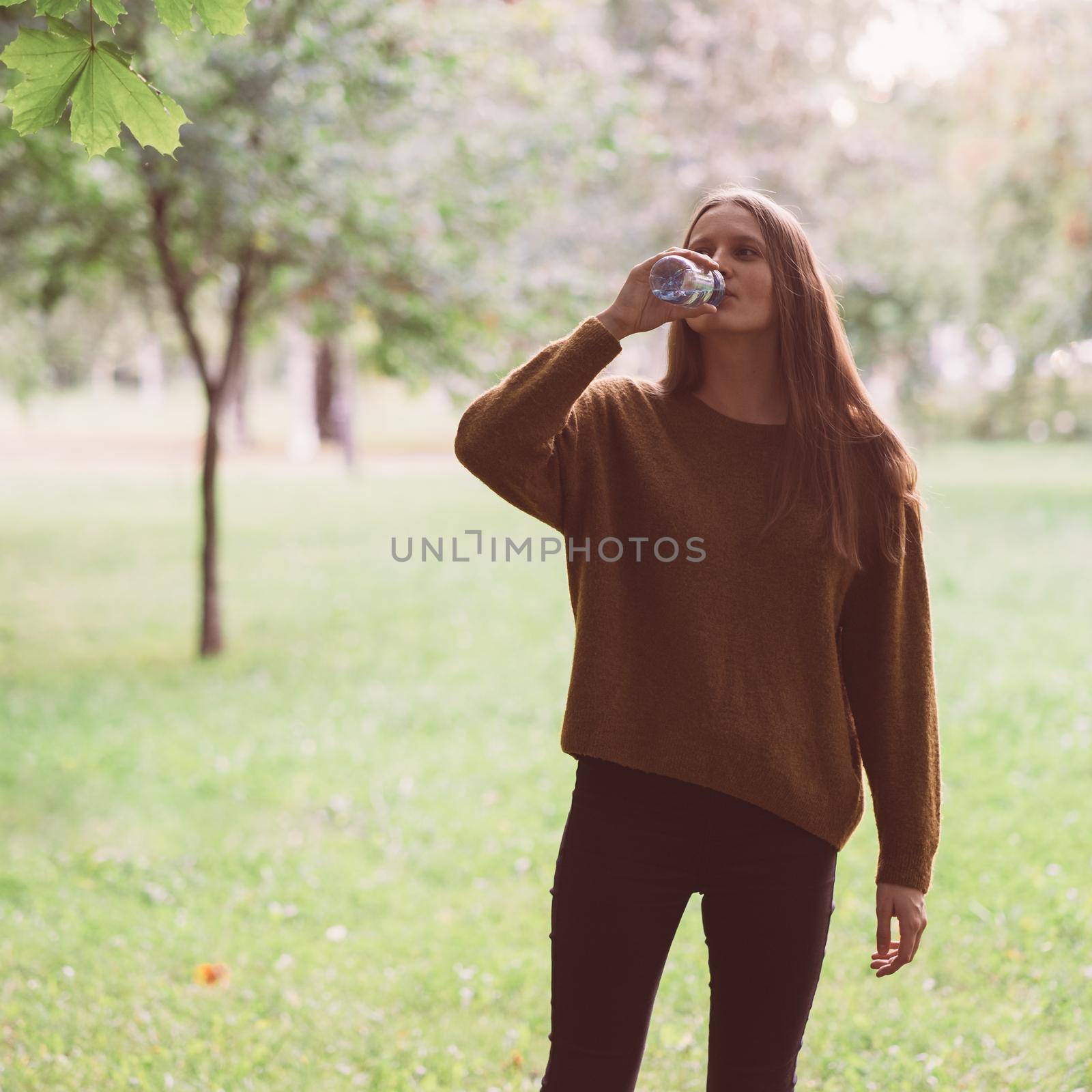 Young beautiful girl drinking water from a plastic bottle on the street in the Park in autumn or winter. A woman with beautiful long thick dark hair quenches her thirst for water on walk by NataBene