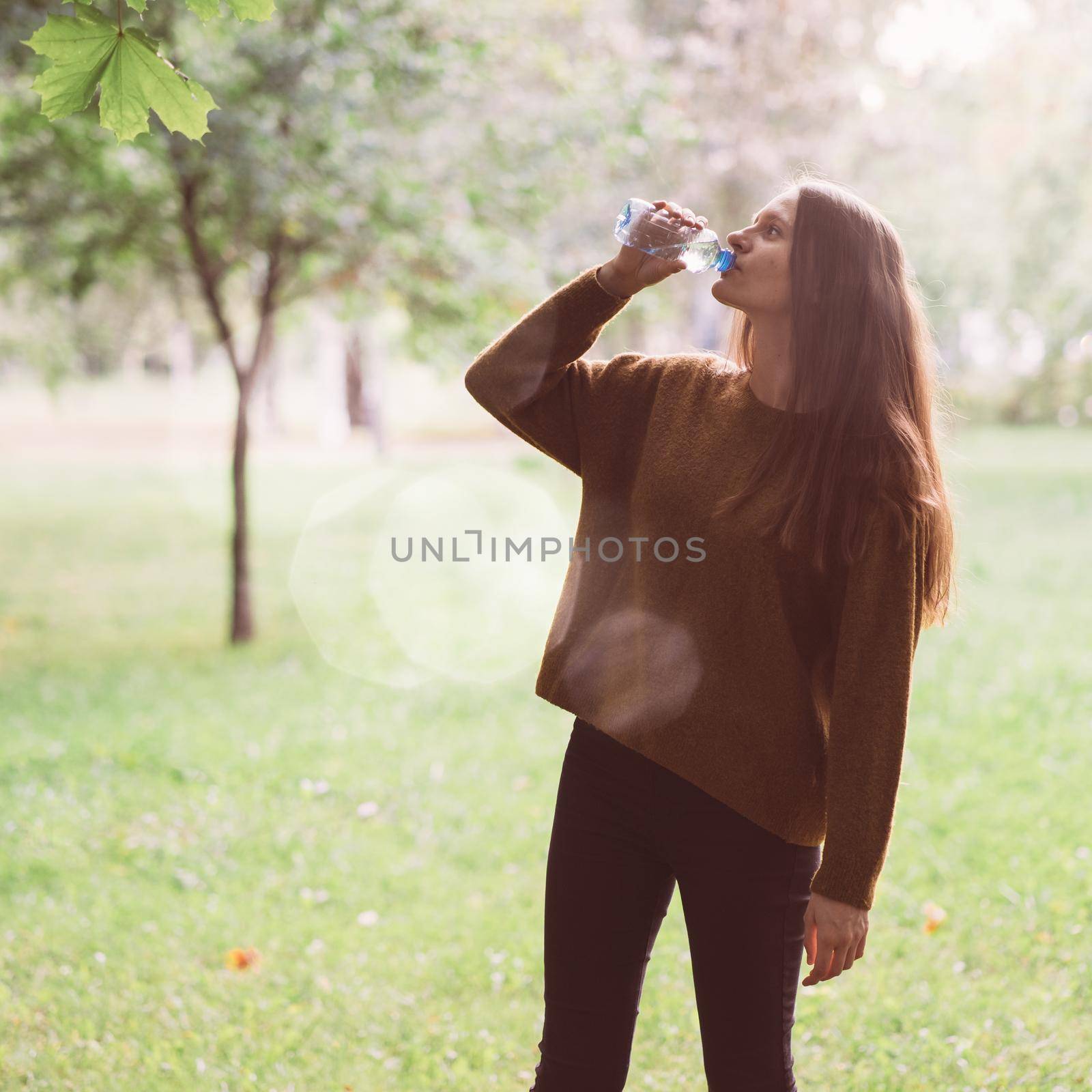 Young beautiful girl drinking water from a plastic bottle on the street in the Park in autumn or winter. A woman in profile with beautiful long thick dark hair quenches her thirst for water on a walk