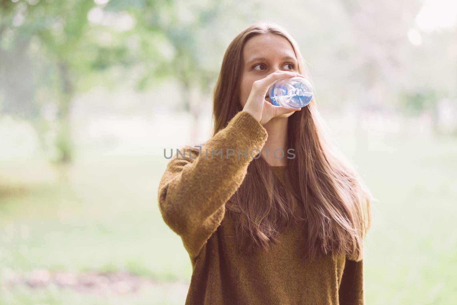 Young beautiful girl drinking water from a plastic bottle on the street in the Park in autumn or winter. A woman with beautiful long thick dark hair quenches her thirst for water on a walk, close up