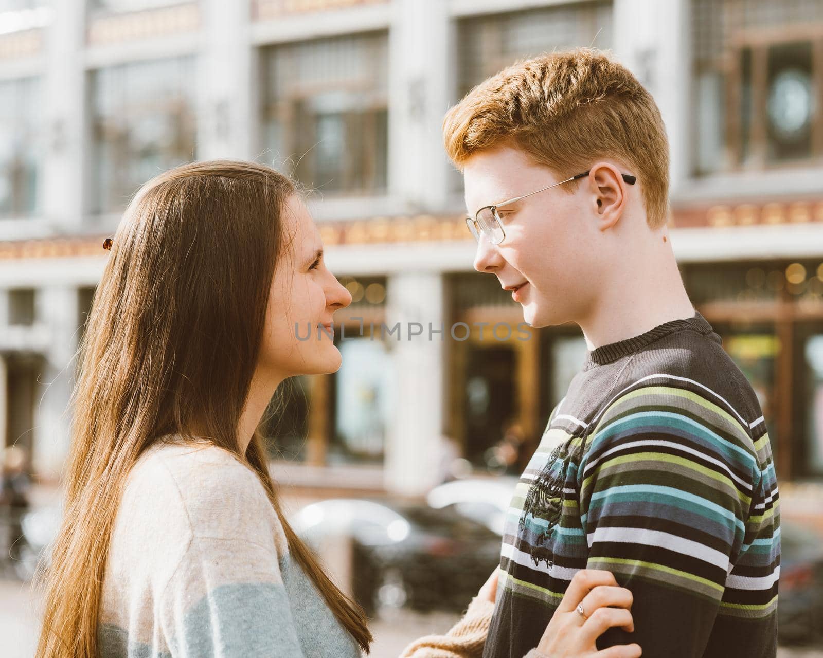Male and female person looking at each other, young couple full of love. The redhead boy looks tenderly at girl and kiss. Concept of teenage love and first kiss, love, relationship. City, waterfront. by NataBene