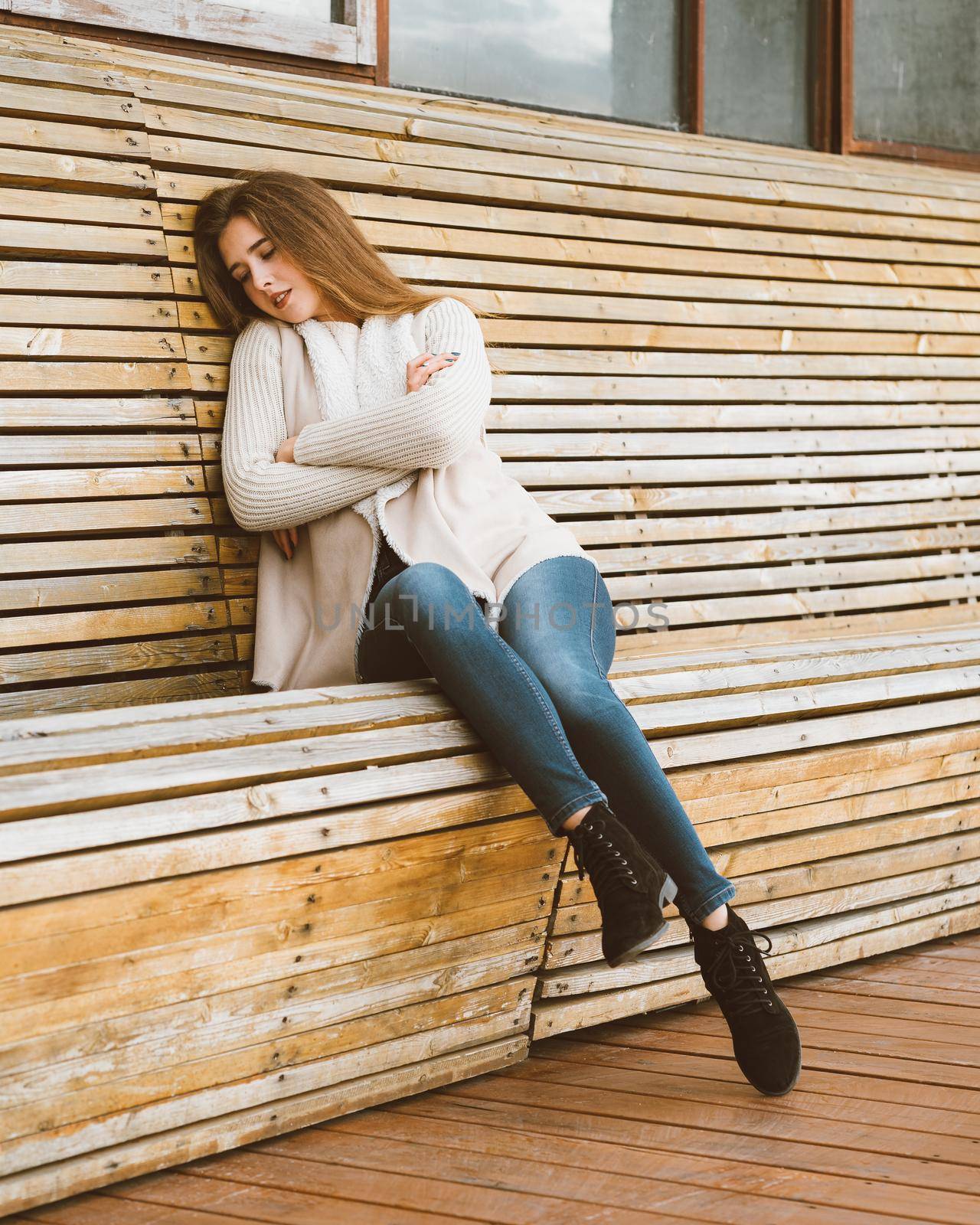 Beautiful young girl with long brown hair sits on wooden bench made of planks and rests, sleeps or dozes in fresh air. Outdoor photo shoot with attractive woman in winter or autumn, vertical