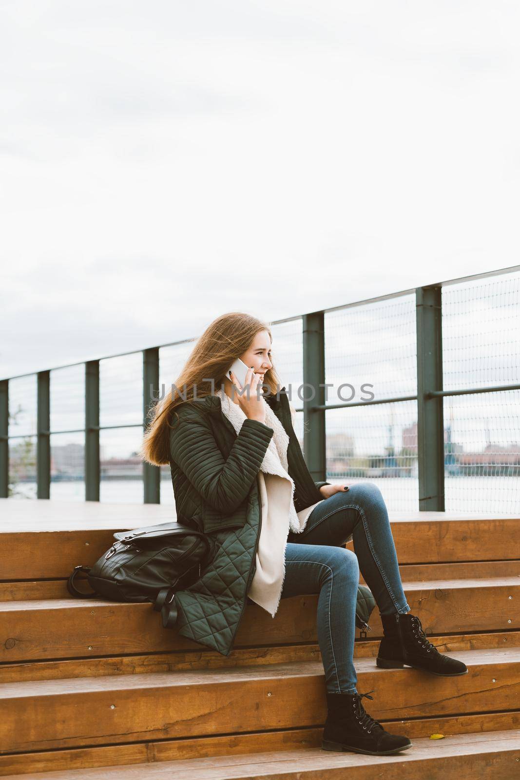 Beautiful woman with long hair talking on phone. Autumn or winter, girl in outdoor jacket sitting on wooden steps by NataBene