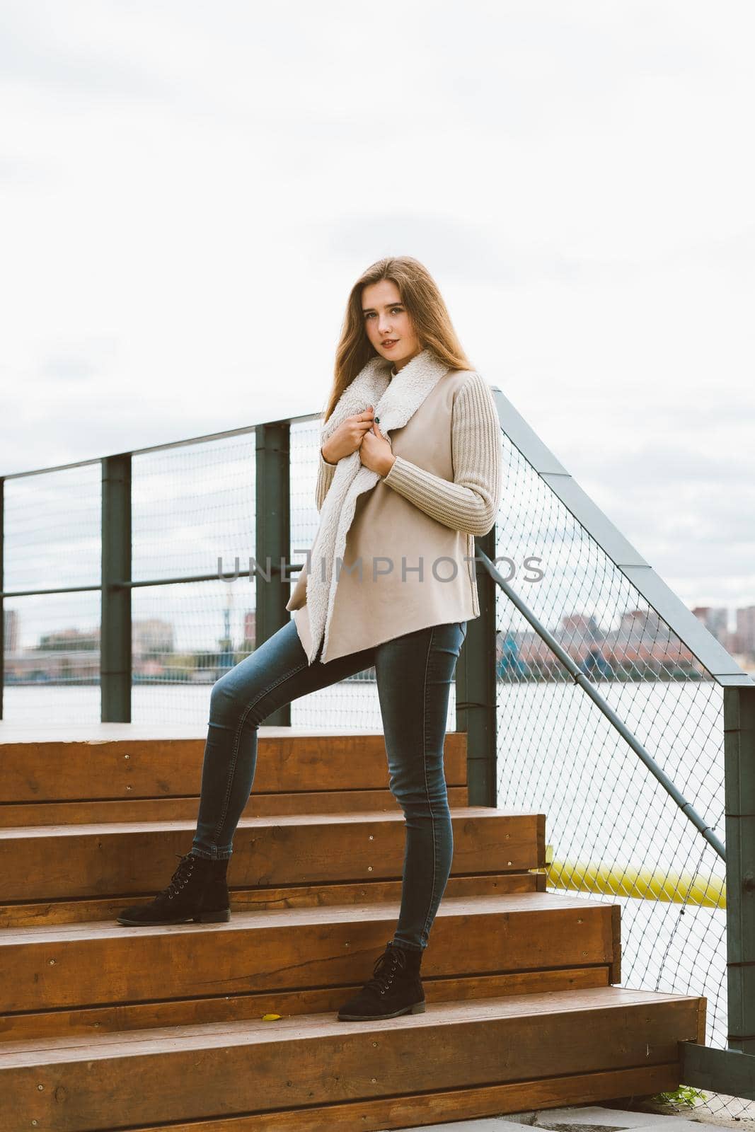 Beautiful young long-haired Caucasian woman stands on steps of wooden platform on waterfront at pier in port, vertical