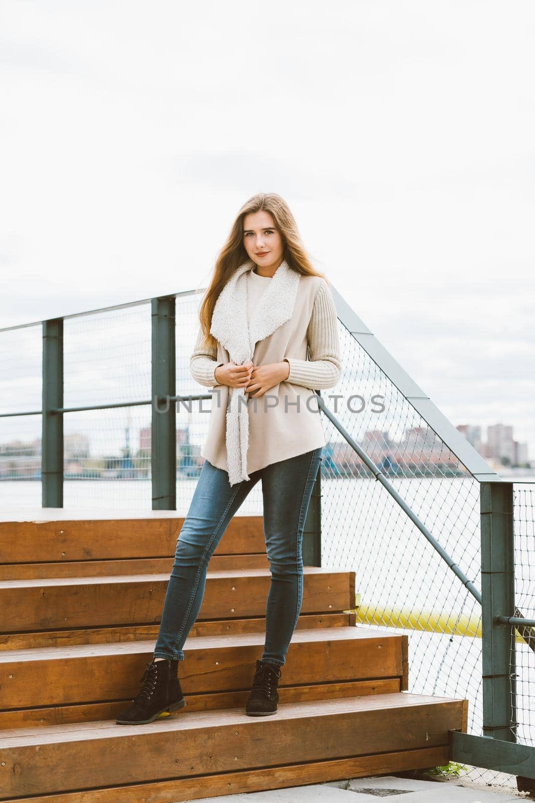 Beautiful young long-haired Caucasian woman stands on steps of wooden platform on waterfront at pier in port. by NataBene