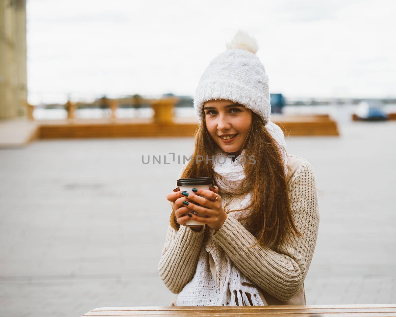 Beautiful young girl looking at camera, holding coffee, tea in reusable plastic mug in autumn, winter. A woman in warm clothes sitting on waterfront on Baltic sea in port, warmed by a hot drink