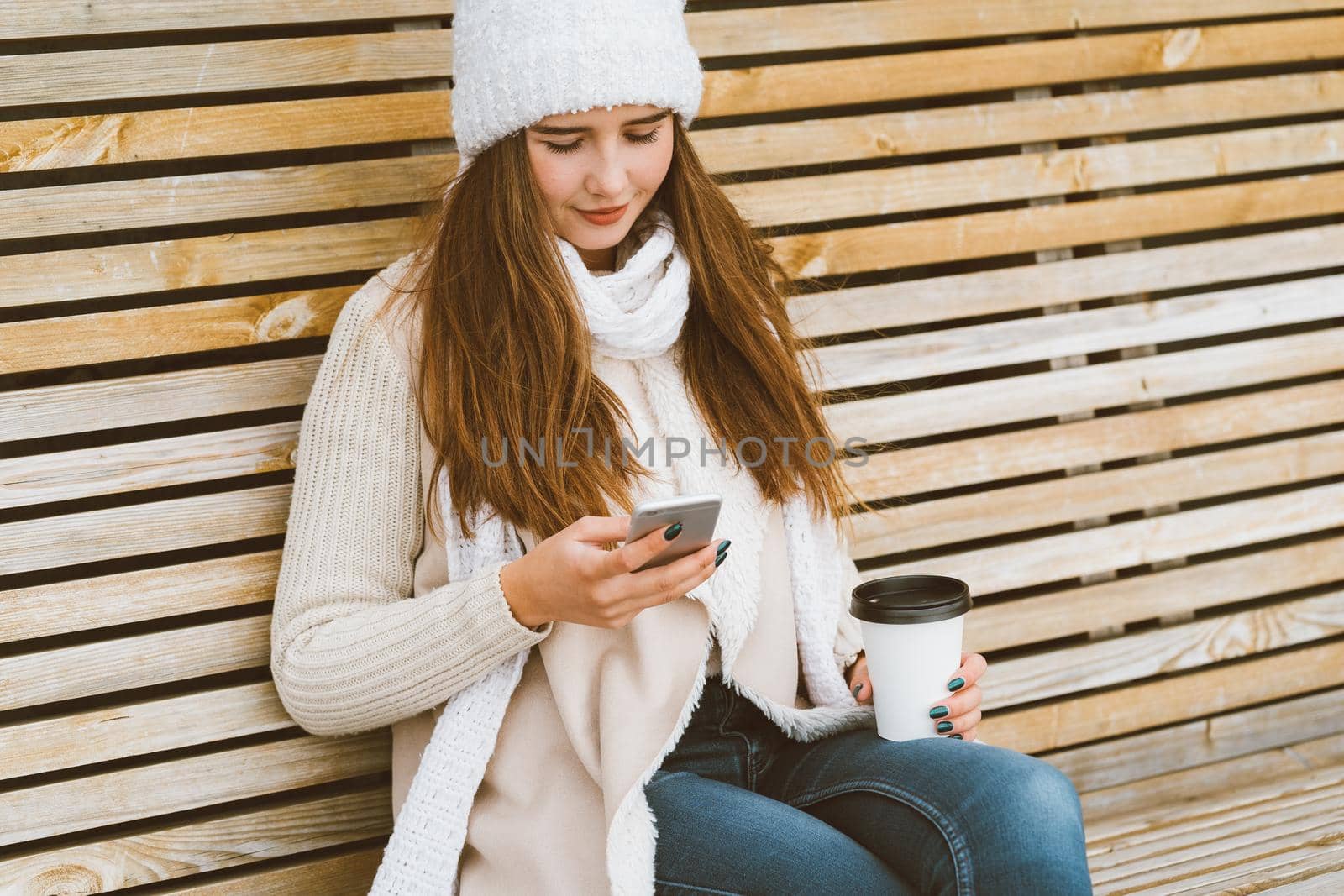Beautiful young girl drinking coffee, tea from plastic mug in autumn, winter and talking on mobile phone. Woman with long hair sitting on bench in autumn or winter, basking in hot drink, copy space by NataBene