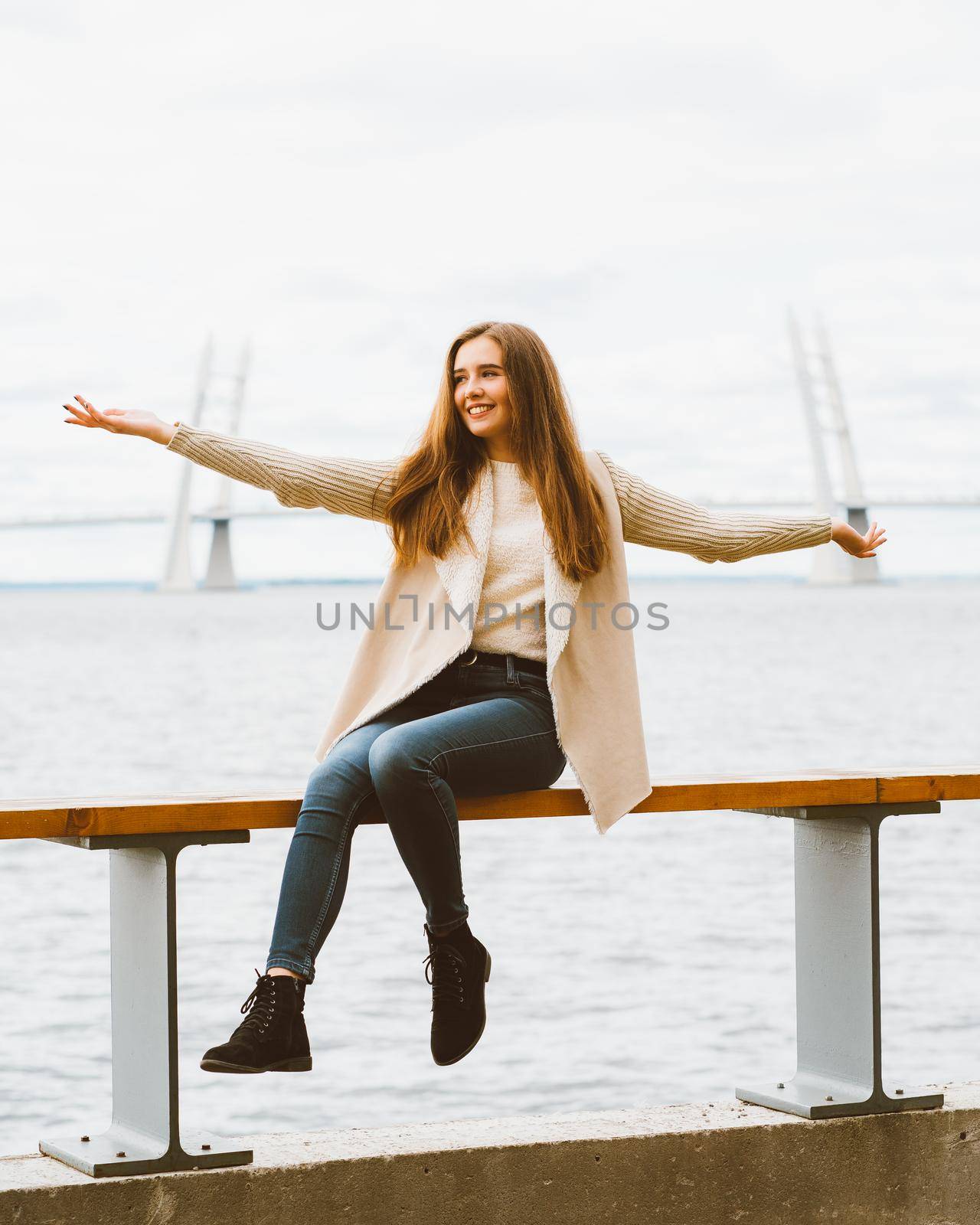 Happy young girl sitting on waterfront at pier in the port, enjoying life and waving his arms. Woman with long hair smiles and enjoys moment by NataBene