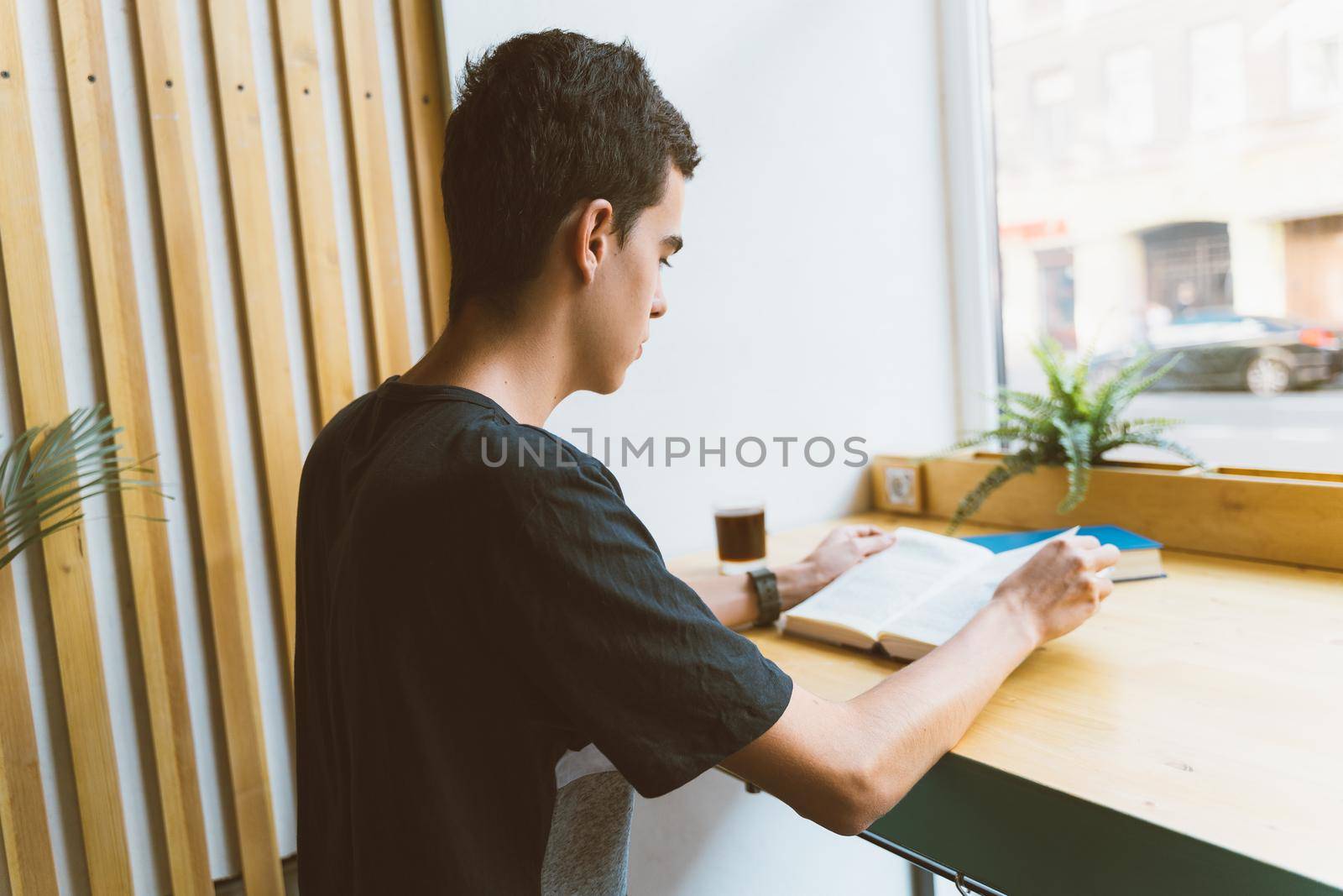 Young boy with black hair sitting by the window in the library reading a big book, teenager in casual clothes in library