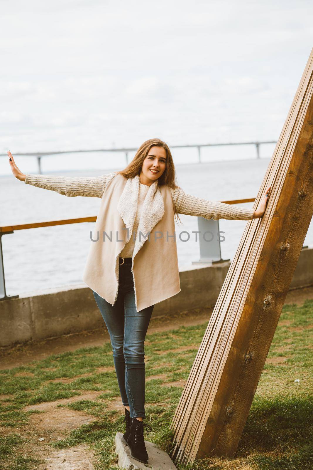 Happy young girl standing on the waterfront at pier in port, enjoying life and waving his arms. Woman with long hair smiles and enjoys moment, vertical