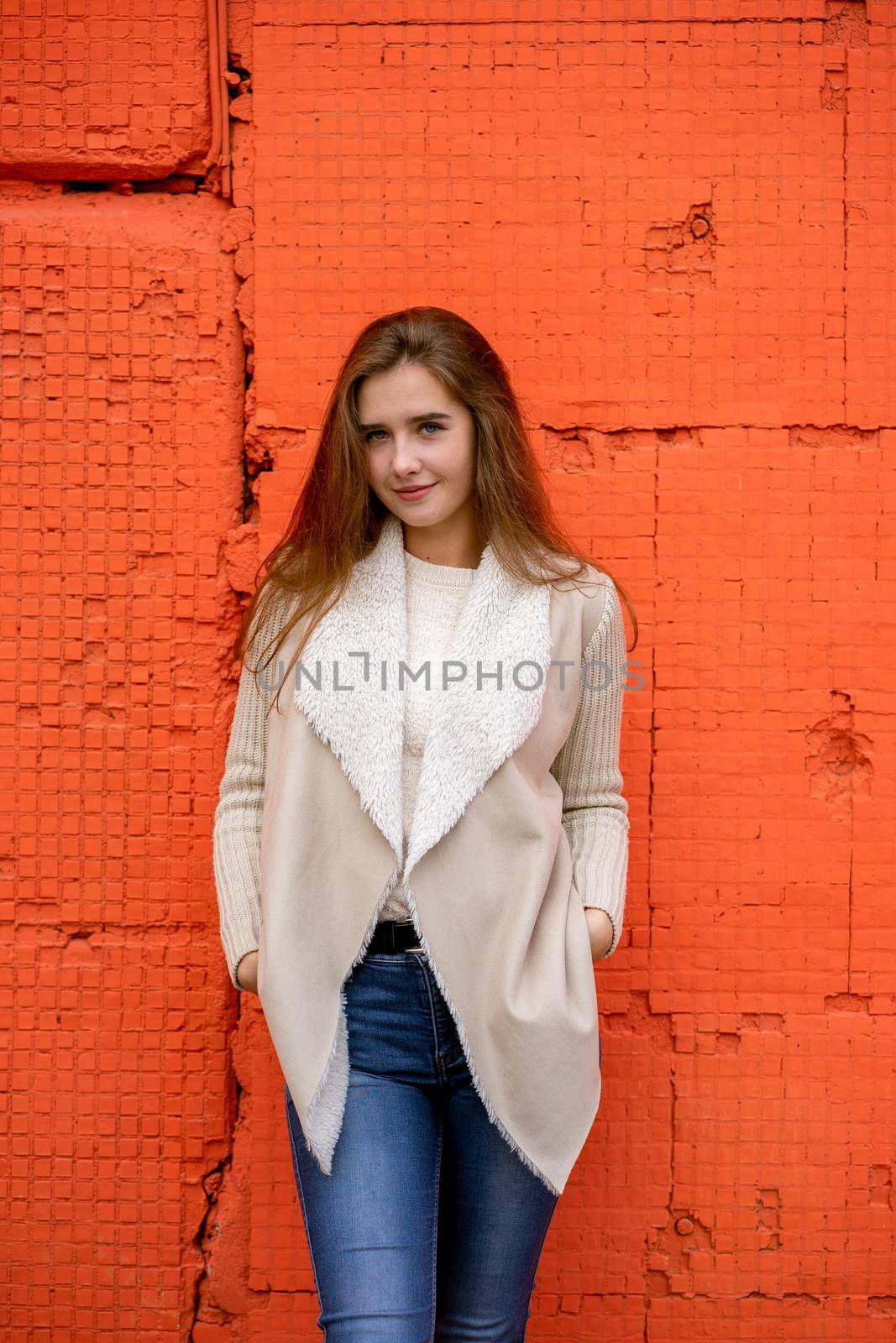 A beautiful girl stands against a bright wall of orange coral color, a woman with long hair in warm clothes on a colorful colored background