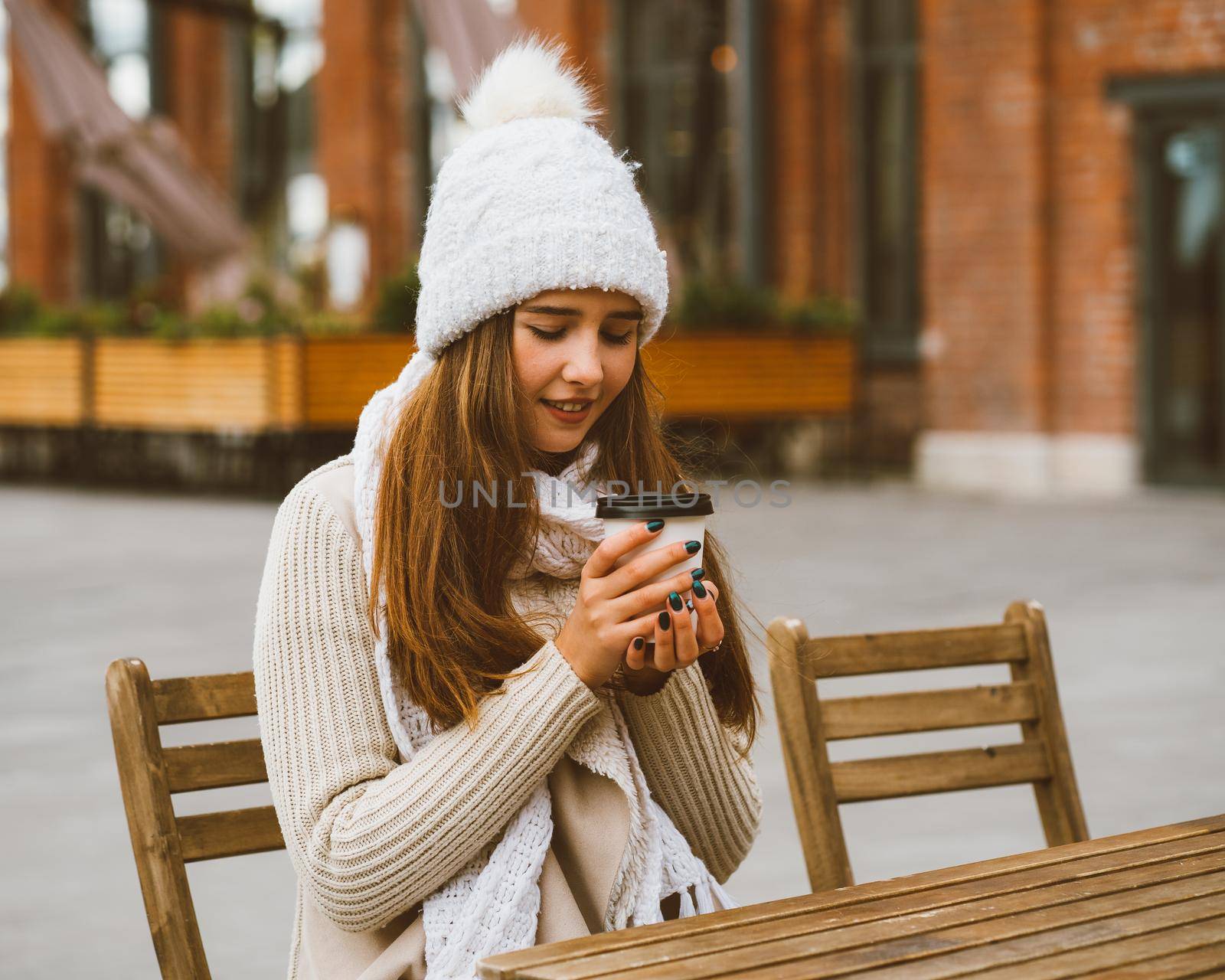 Beautiful young girl drinking coffee, tea from plastic mug in autumn, winter. A woman with long hair in warm clothes sitting in street cafe, warmed by hot drink by NataBene