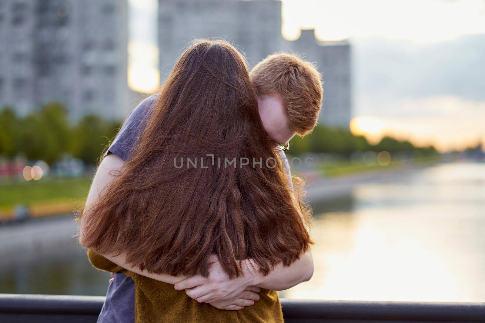 Girl with long thick dark hear embracing redhead boy on bridge, teen love at sunset
