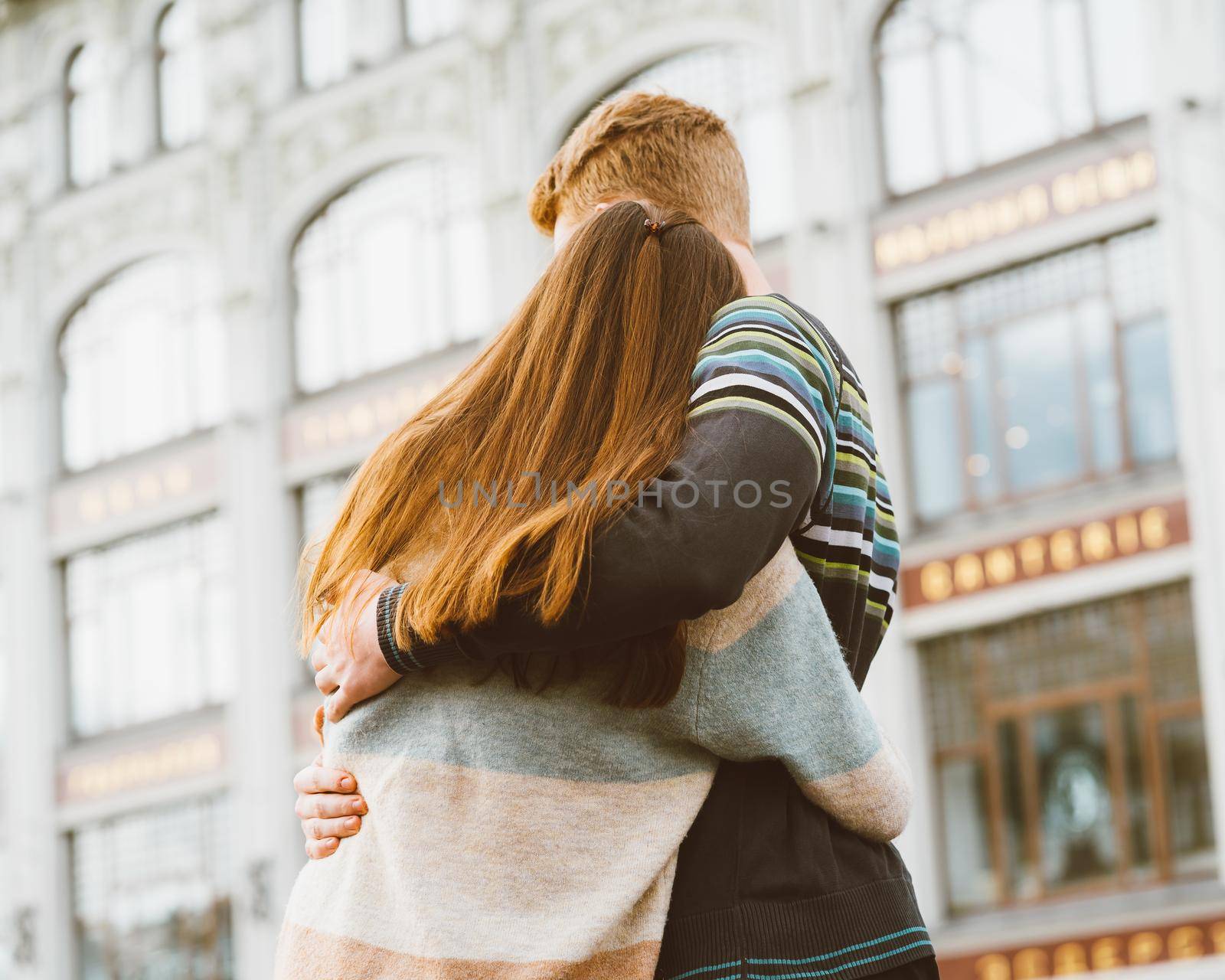 Girl with long thick dark hair holding hands redhead boy in blue t-shirt on bridge, teen love at evening. Boy looks tenderly at a girl, young couple. Concept of teenage love and first kiss, sincere feelings of man and woman, city, waterfront. Close up