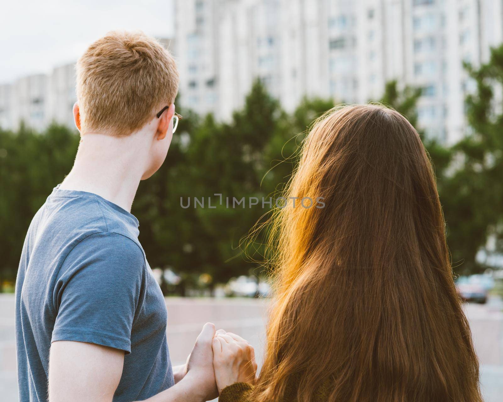 Girl with long thick dark hair holding hands redhead boy in blue t-shirt on bridge, teen love at evening. Boy looks tenderly at a girl, young couple. Concept of teenage love and first kiss, sincere feelings of man and woman, city, waterfront. Close up