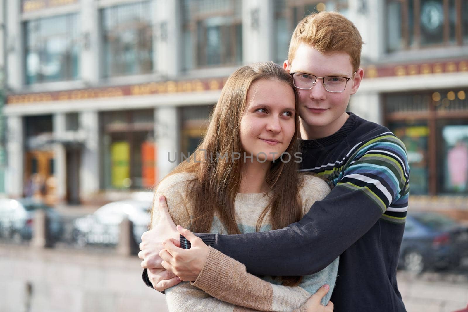 portrait of happy couple embracing in downtown, red-haired man with glasses looking straight, woman with long hair looking away, copy space