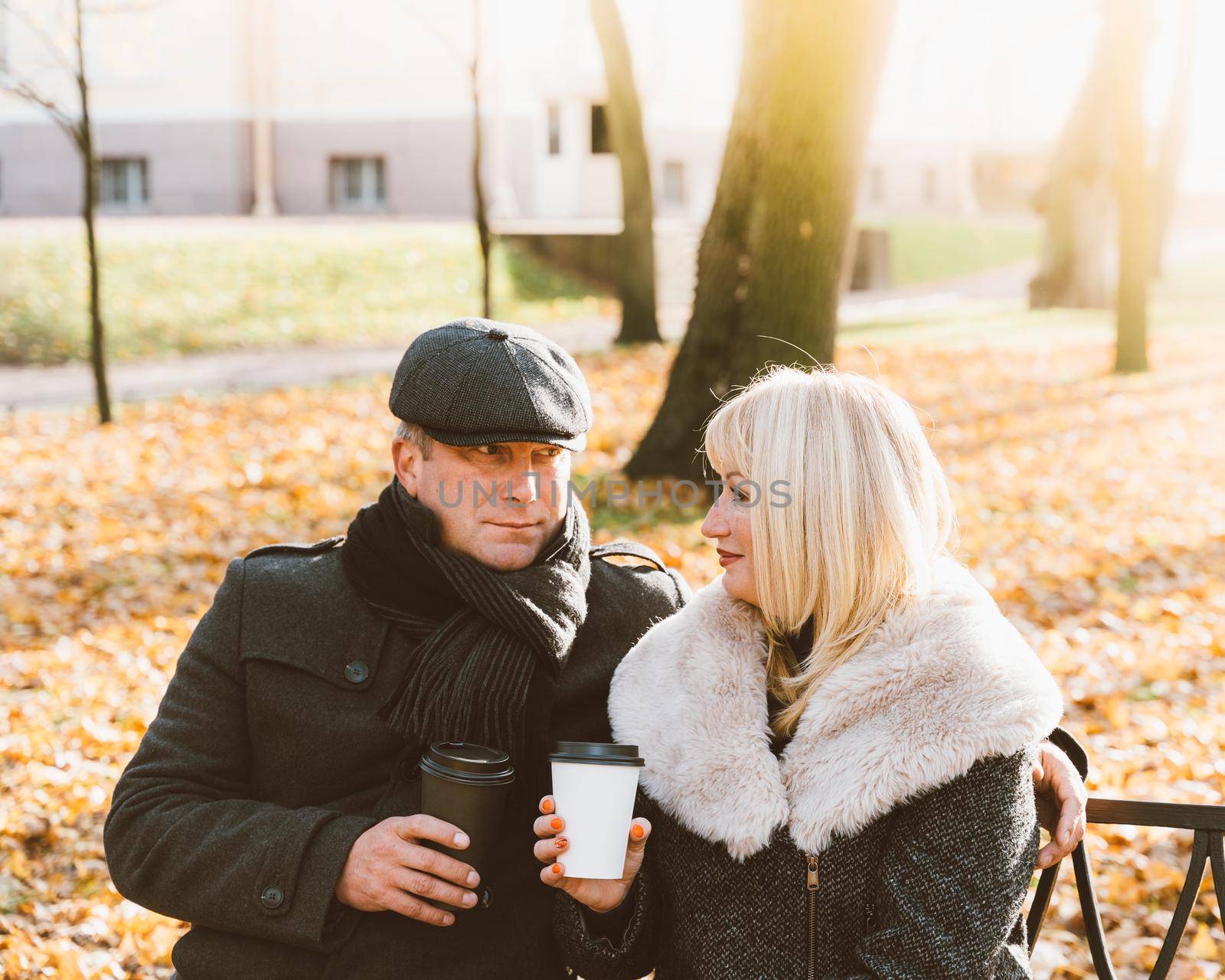 Happy blonde mature woman and handsome middle-aged brunette man sit on bench and drink coffee from a refillable cup. A loving couple of 45-50 years old walks in autumn or winter park in warm clothes