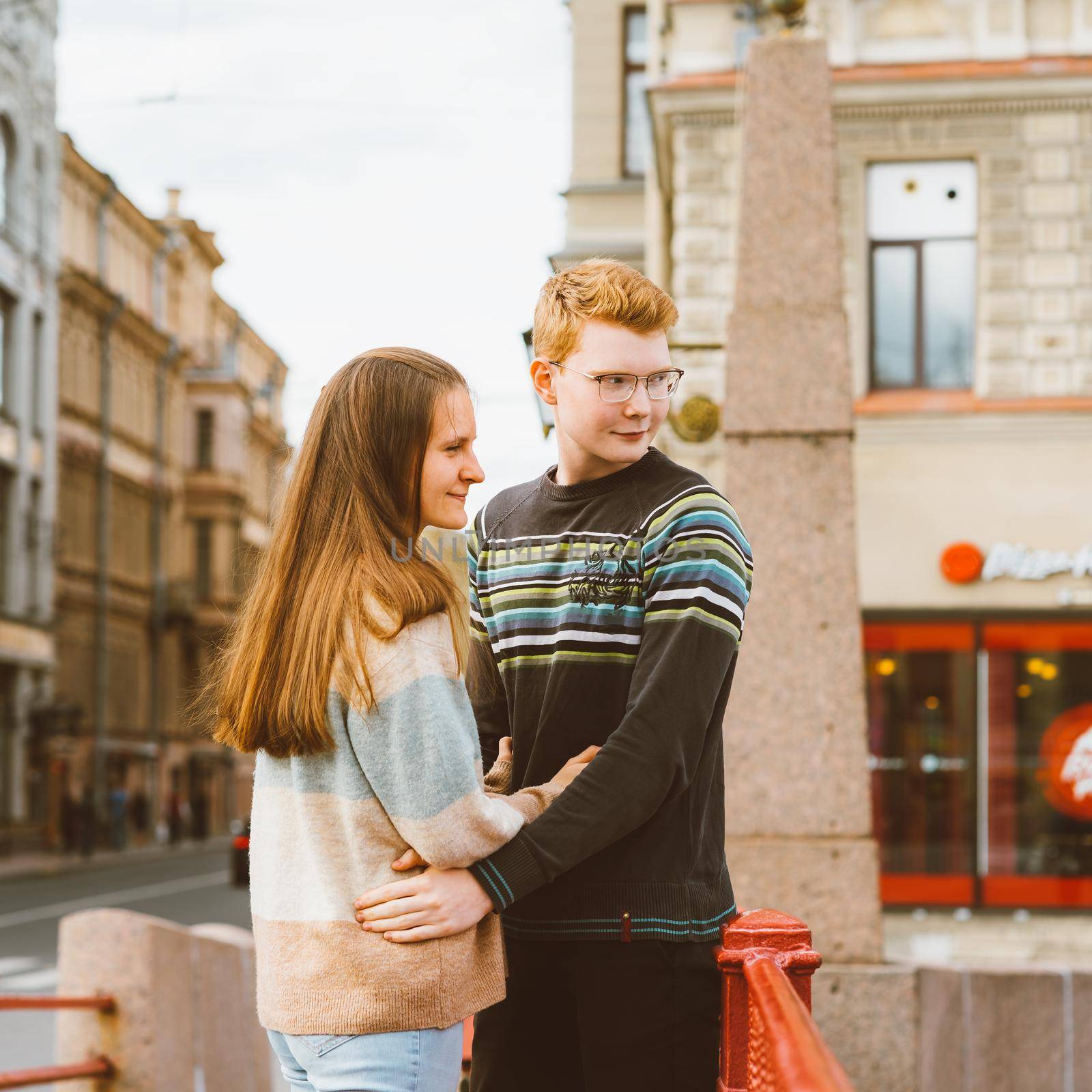 Girl with long thick dark hear embracing redhead boy in blue t-shirt on bridge, young couple. Concept of teenage love. they look away, saw something. City, waterfront. by NataBene