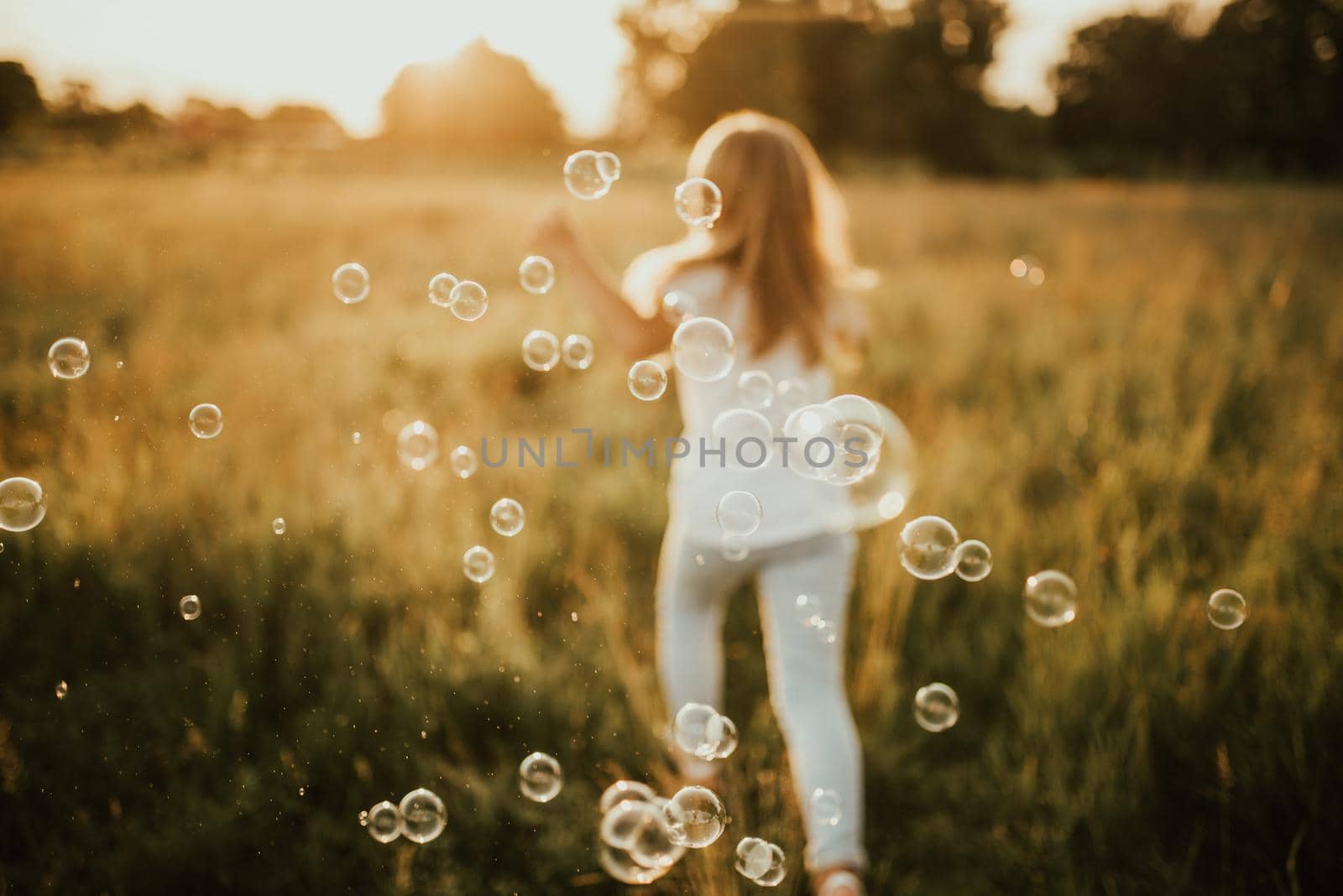 a little blonde girl dressed in light-colored clothes and a white T-shirt runs away from soap bubble. Along the tall green grass towards the setting sun. Warm summer sunny weather at sunset.