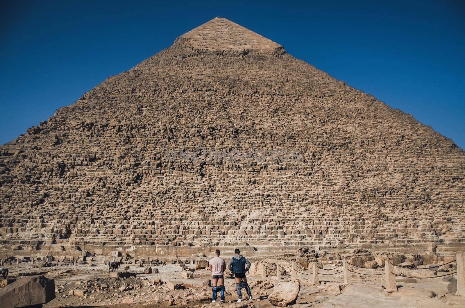 Two men stand in front of old pyramid with their pants down. by AndriiDrachuk