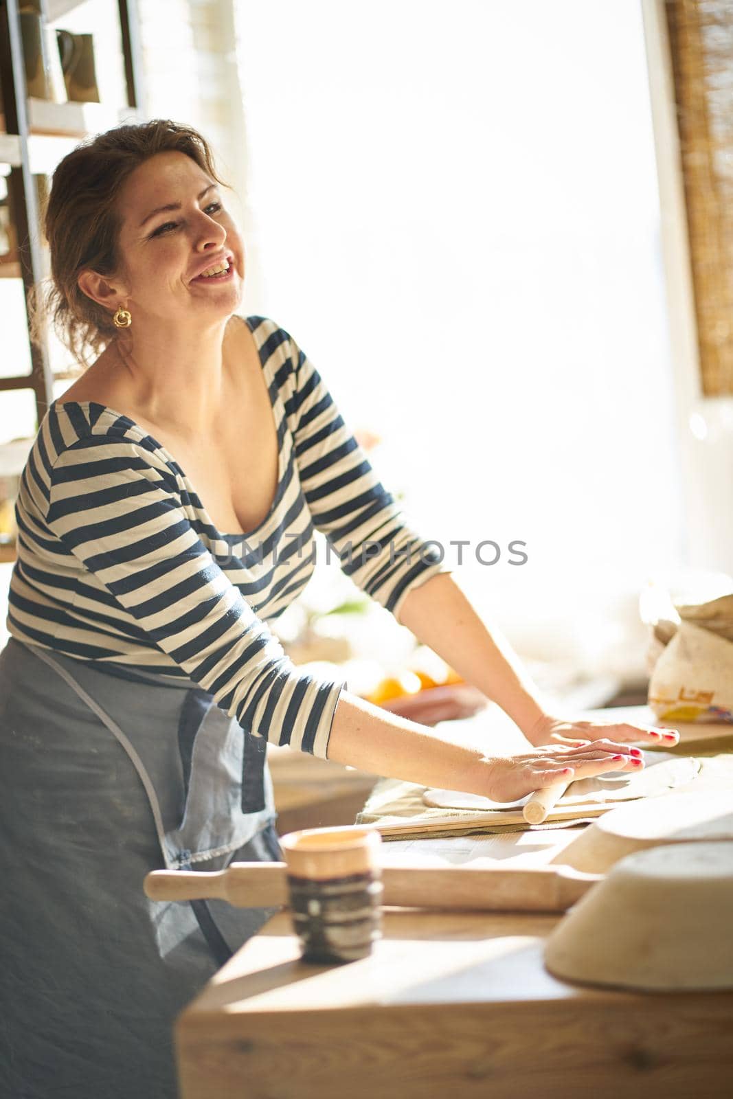 Beautiful happy woman making ceramic ware in workplace, laughing, smiling in sun light. Concept for woman in freelance, business, hobby