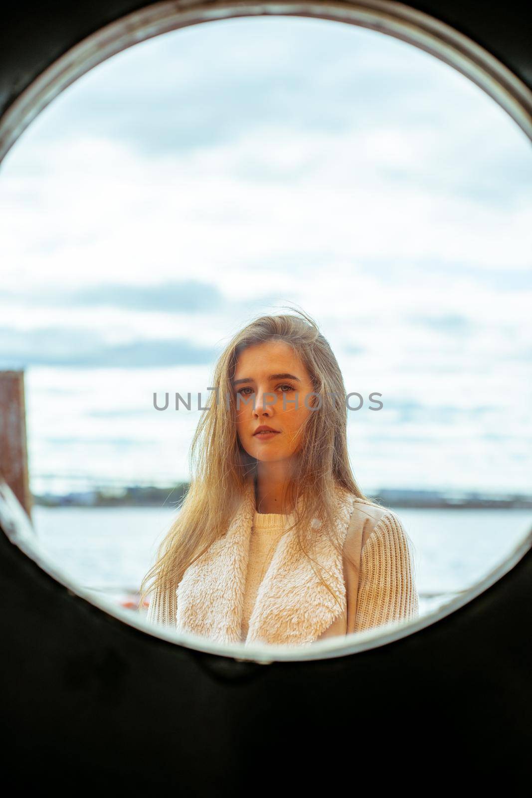 The face of young girl in round frame window on background of sea, ocean, on waterfront. Portrait in a circle, backlit, vertical