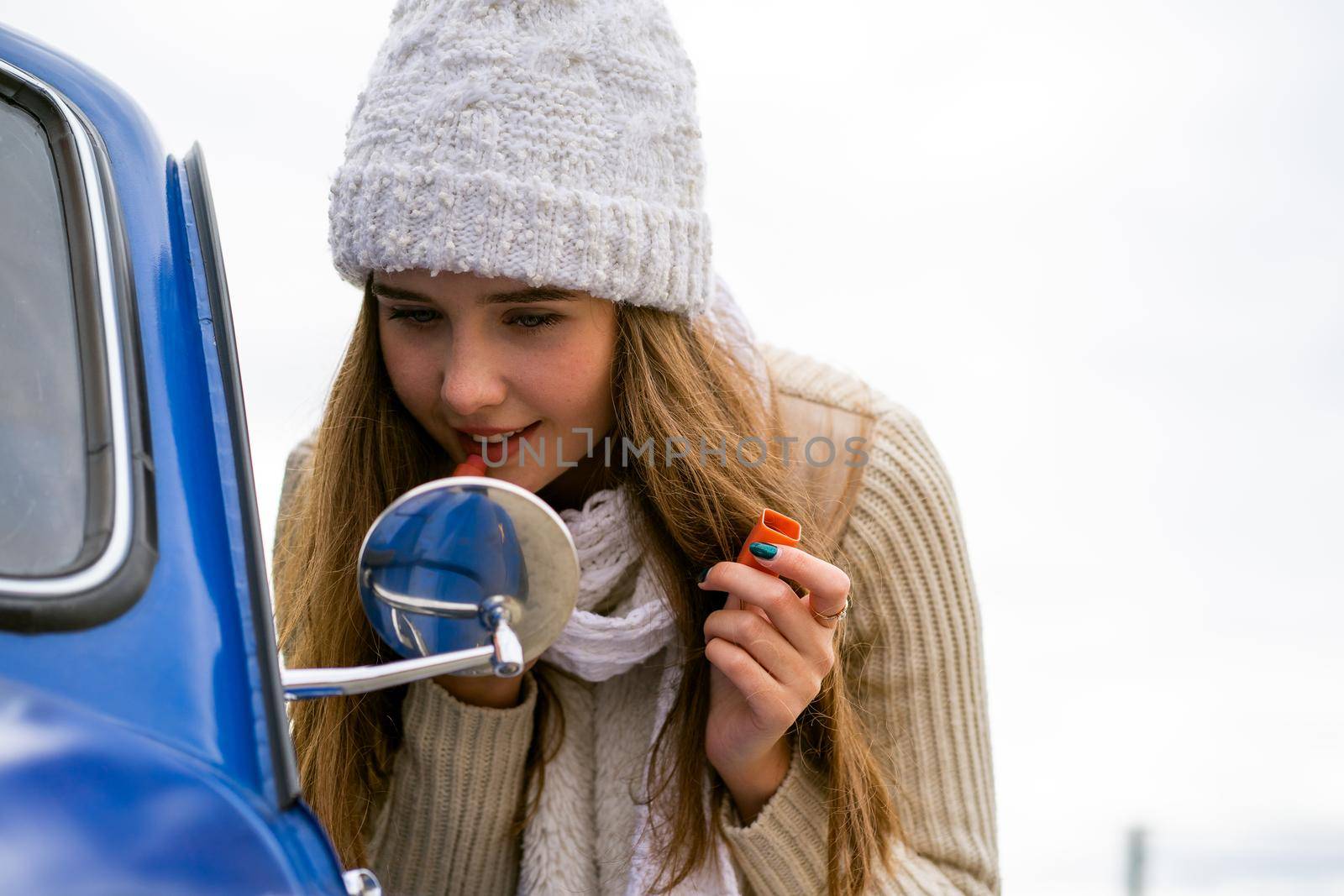 Beautiful young girl looks in the car mirror and make up her lips. Woman with long hair in hat and scarf next to blue retro car in autumn or winter, copy space